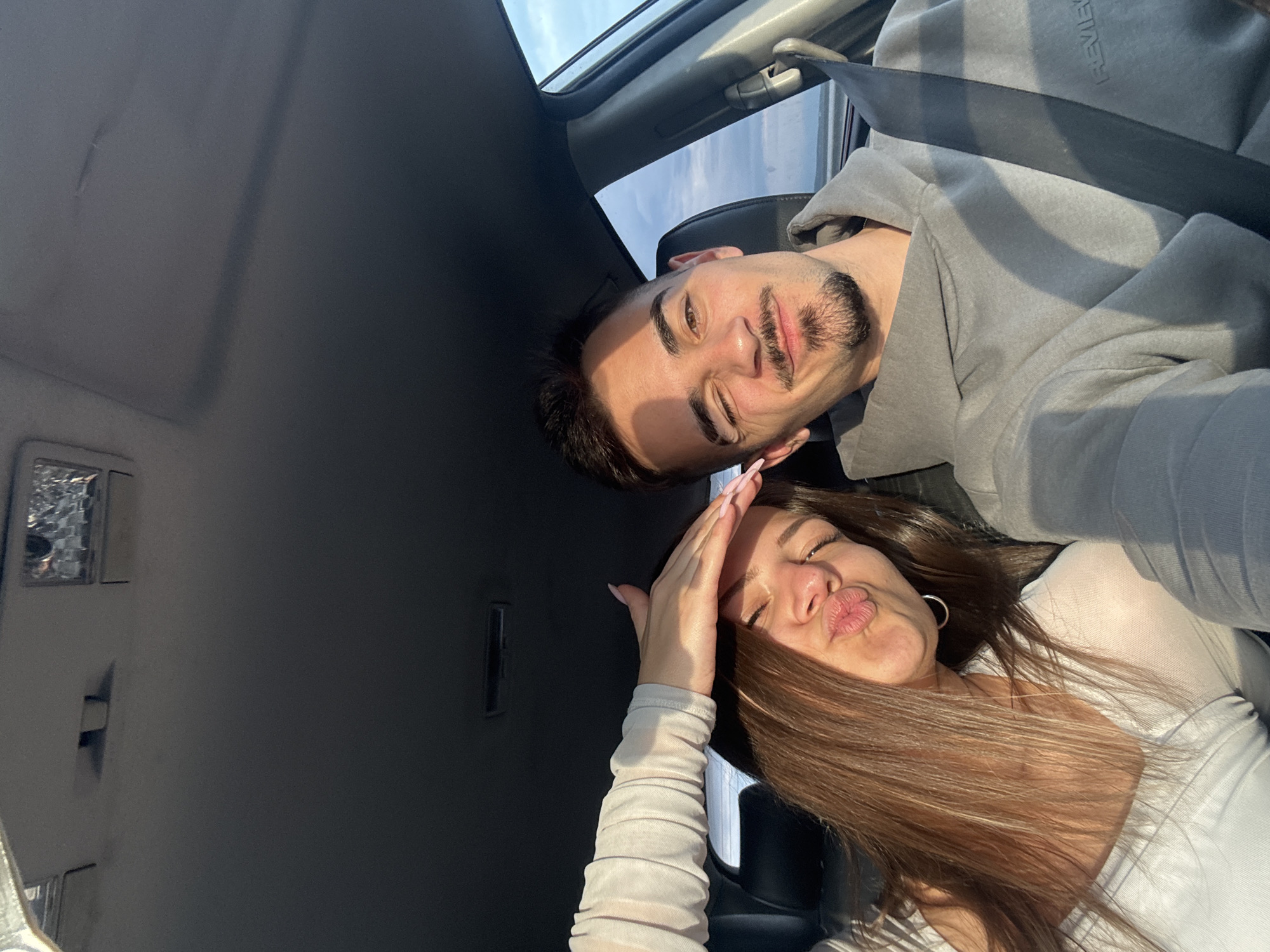 A man and woman are posing for a selfie inside a car. The man has dark hair and a beard, wearing a gray hoodie and a seatbelt. The woman has long brown hair, wearing a white top, and is making a kissing face while holding her hand up to her forehead. The car's interior is gray. The location is unknown, and there is no evidence to suggest the city of Chokoba, Bulgaria.