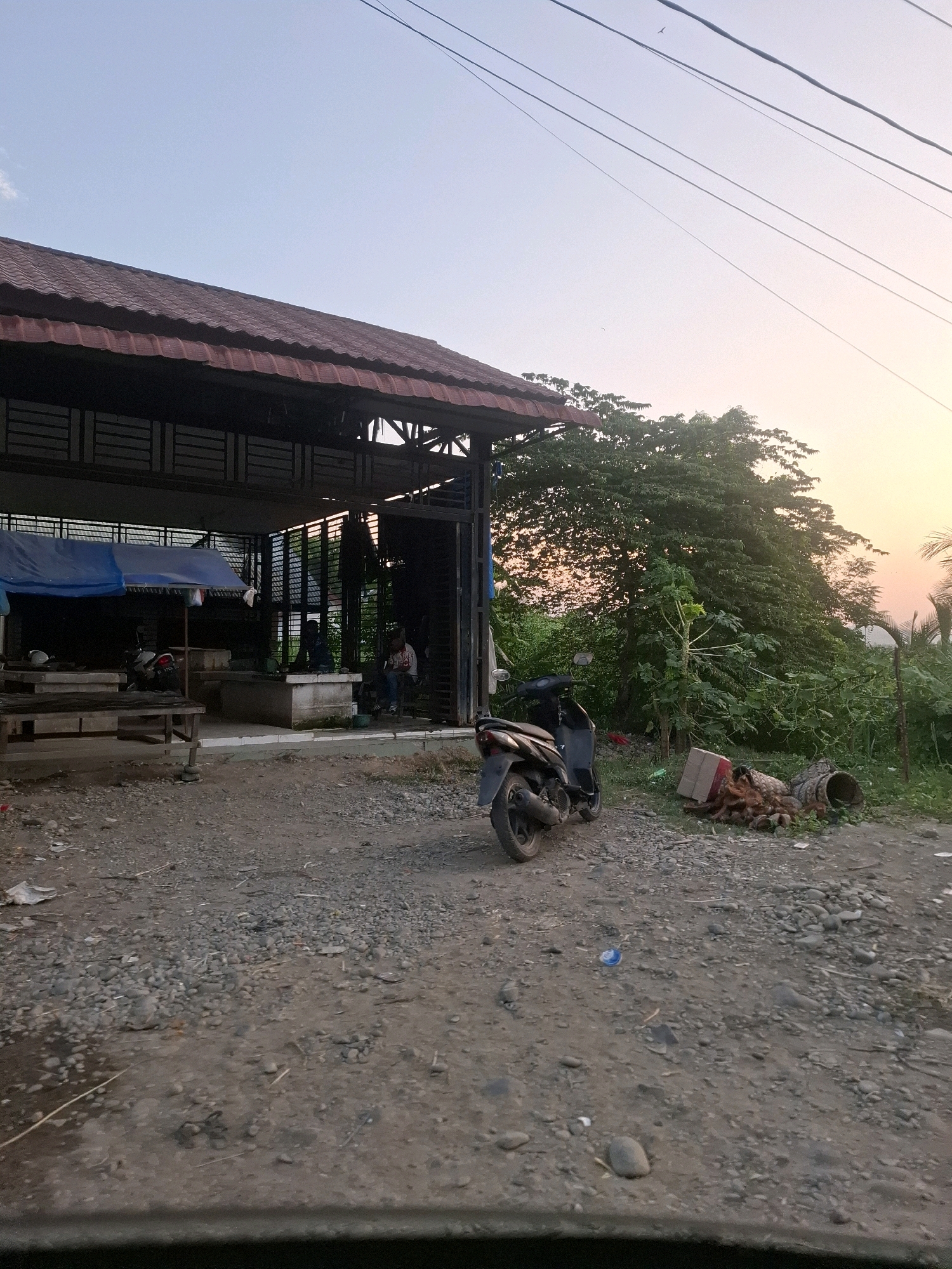 The image depicts an outdoor scene at dusk in what appears to be a semi-rural or underdeveloped area, possibly in Keudejangkabuya, Indonesia. The foreground is a dirt and gravel ground littered with small rocks and some debris, including a blue plastic cap. To the left, a covered structure with a reddish-brown tiled roof is visible. Beneath this structure, a blue tarp is partially covering some items, and a motorcycle is parked. Inside the structure, there are several concrete platforms or tables, and at least two individuals are seated, one appearing to be wearing a dark shirt and another in a patterned shirt and blue jeans.
To the right of the structure, a dark-colored scooter is parked on the gravel. Further to the right, a large, leafy tree dominates the midground, with lush green foliage. In front of the tree, a pile of debris is present, including a cardboard box, what appear to be coconut husks or similar plant matter, and a cylindrical object. The background shows more dense vegetation and trees, with the sky transitioning from a pale blue at the top to a warm orange-yellow hue near the horizon, indicating sunset or sunrise. Several power lines stretch across the upper portion of the image. The overall impression is one of a casual, outdoor gathering or market area at the end of the day.