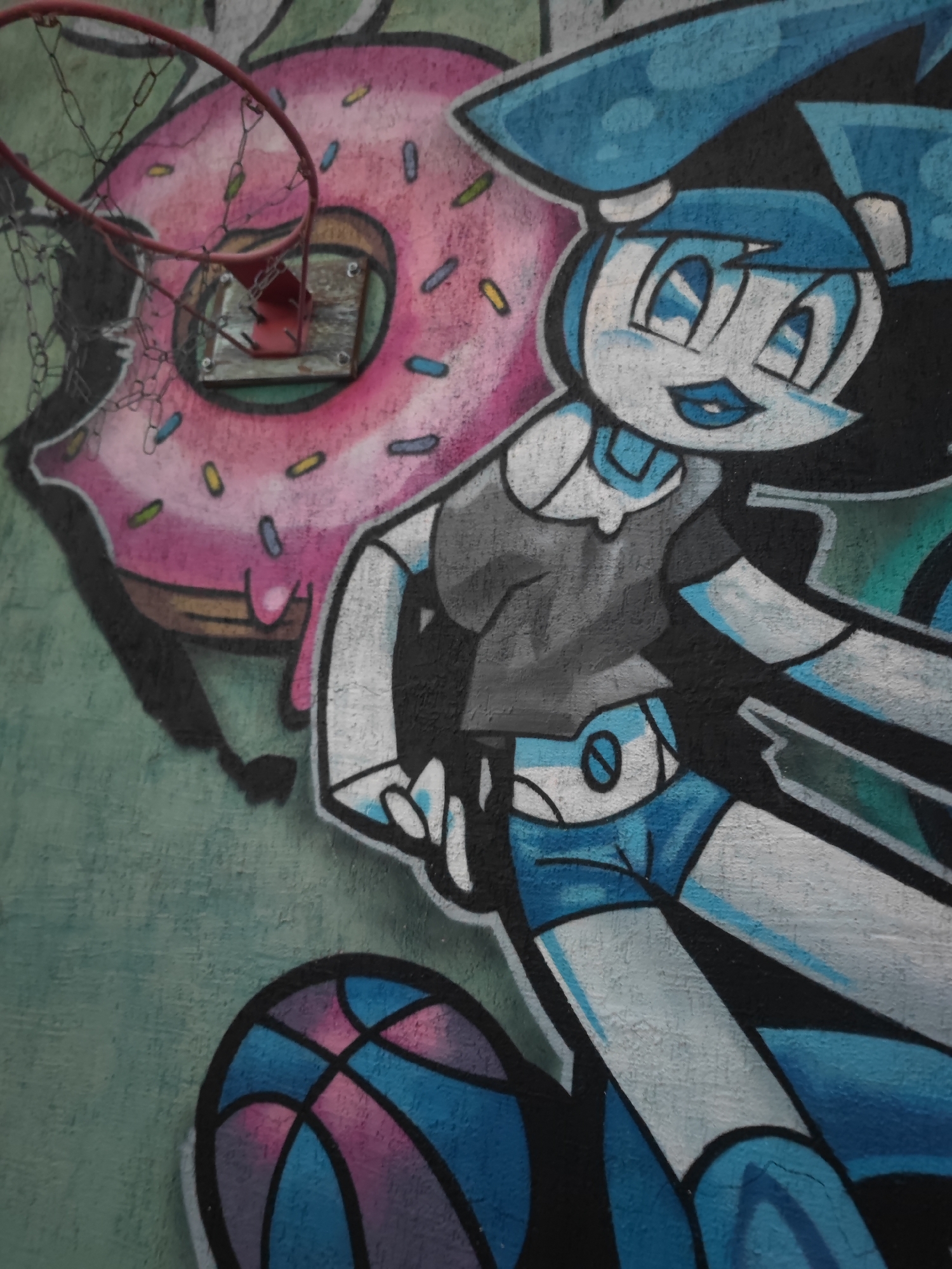 The image is a photograph of graffiti on a wall. The graffiti depicts a cartoon character with blue hair and large blue eyes, wearing a gray shirt and blue shorts. To the left of the character is a large pink donut with sprinkles. A basketball hoop is mounted above the donut. The graffiti is colorful and appears to be on an exterior wall, likely in an urban or semi-urban setting. The specific location is identified as Tuzdybastau, Kazakhstan. The wall surface is textured and appears weathered. The graffiti art style is characteristic of street art.