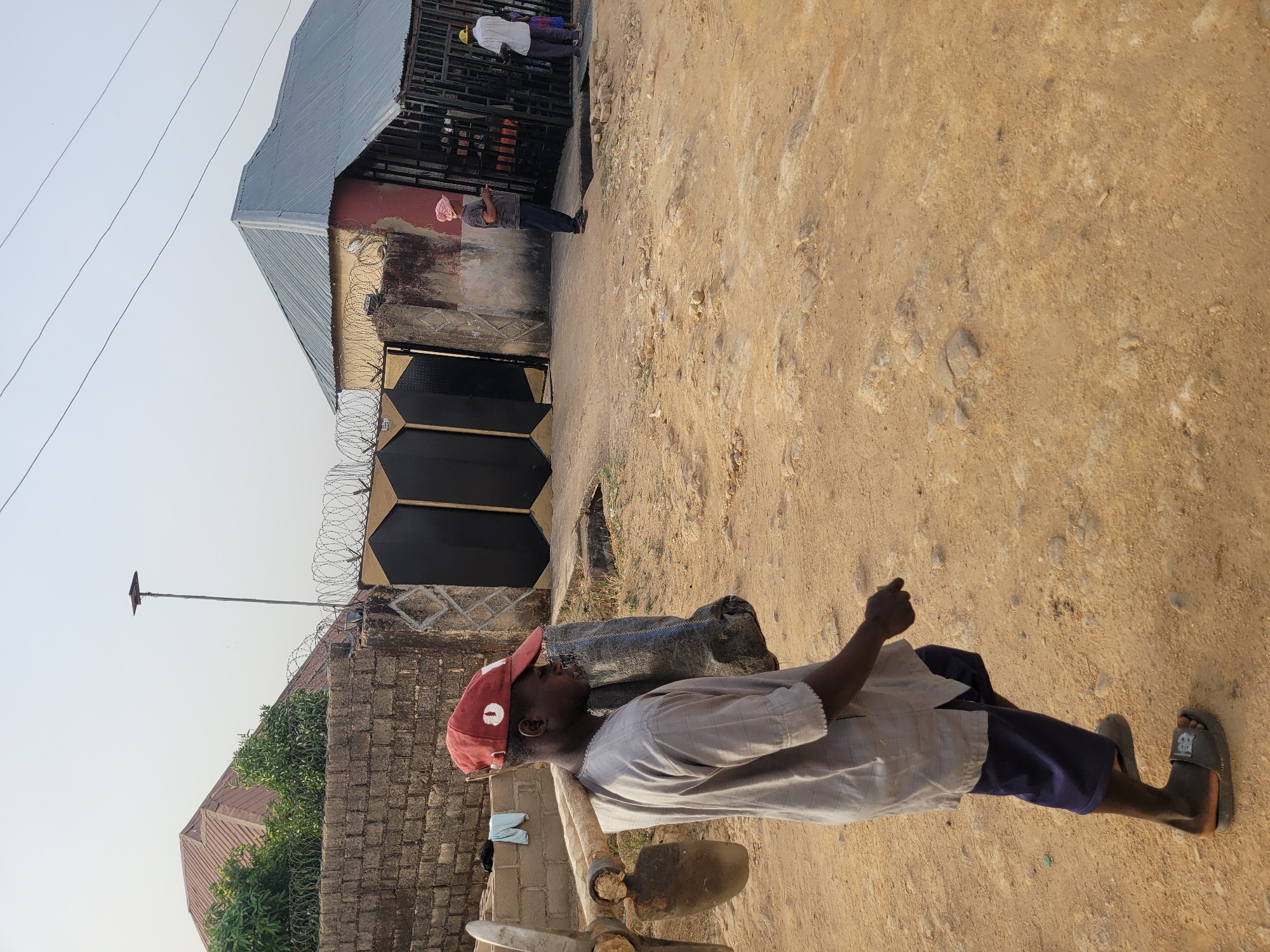 A daytime shot, taken in Barakin Akawo, Nigeria, features an unpaved, dirt road. An African man with a red baseball cap stands in the foreground facing left, carrying a tool with a shovel head. A building is visible behind him, featuring a black gate secured with barbed wire. Two figures appear in the background, standing near the building's entrance.