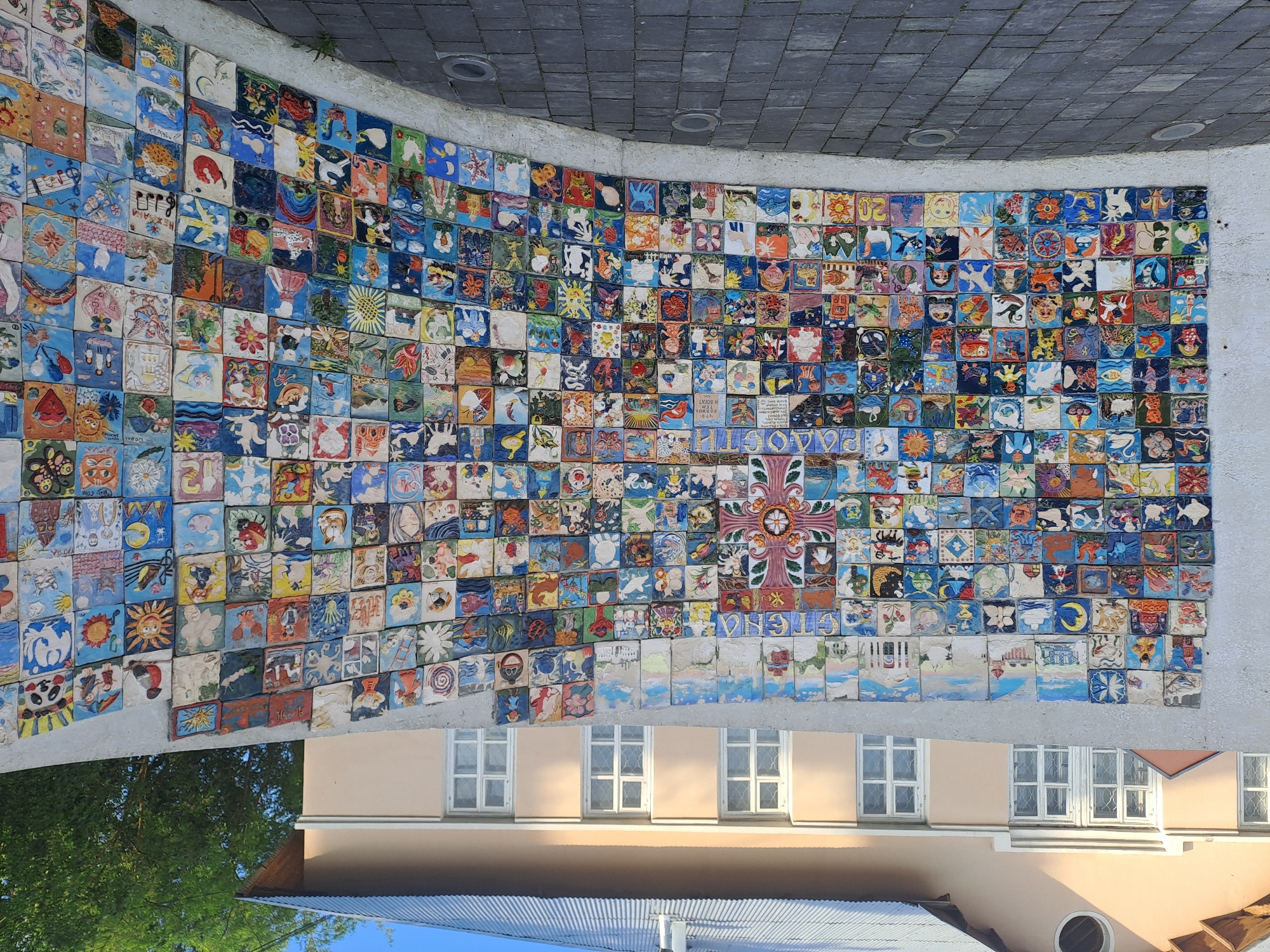 The image shows a curved wall covered with numerous small, colorful tiles, each featuring a unique design. A large, ornate cross made of similar tiles is centrally located on the wall. Above the wall is a paved surface, and below, a building with windows is visible. Lush green trees are seen in the lower left corner. The image is taken in Elektrostal, Russia.