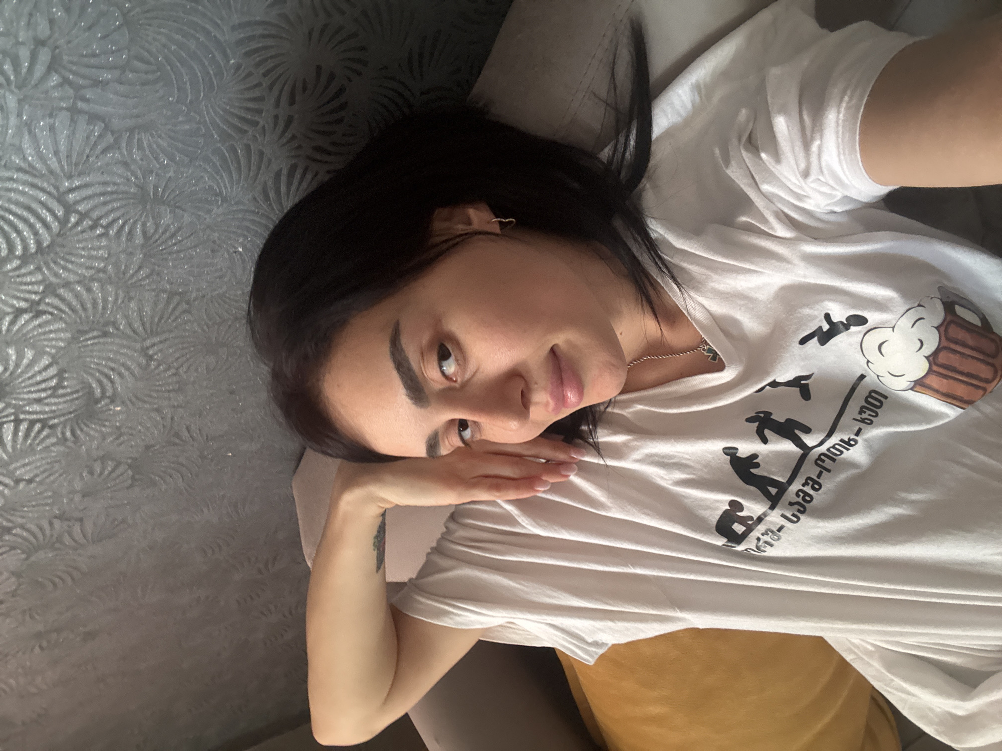 A medium shot depicts a woman lying on a couch with a patterned grey wall in the background. She has dark hair and is wearing a white t-shirt with a black design of people walking up a hill and a beer mug. The design includes Georgian text, suggesting a connection to the city of Tbilisi, Georgia. The woman is resting her head on her hand, looking directly at the camera. A small heart-shaped earring is visible in her left ear. A gold chain necklace is around her neck. A tattoo is visible on her right arm. The couch appears to be grey and mustard yellow.