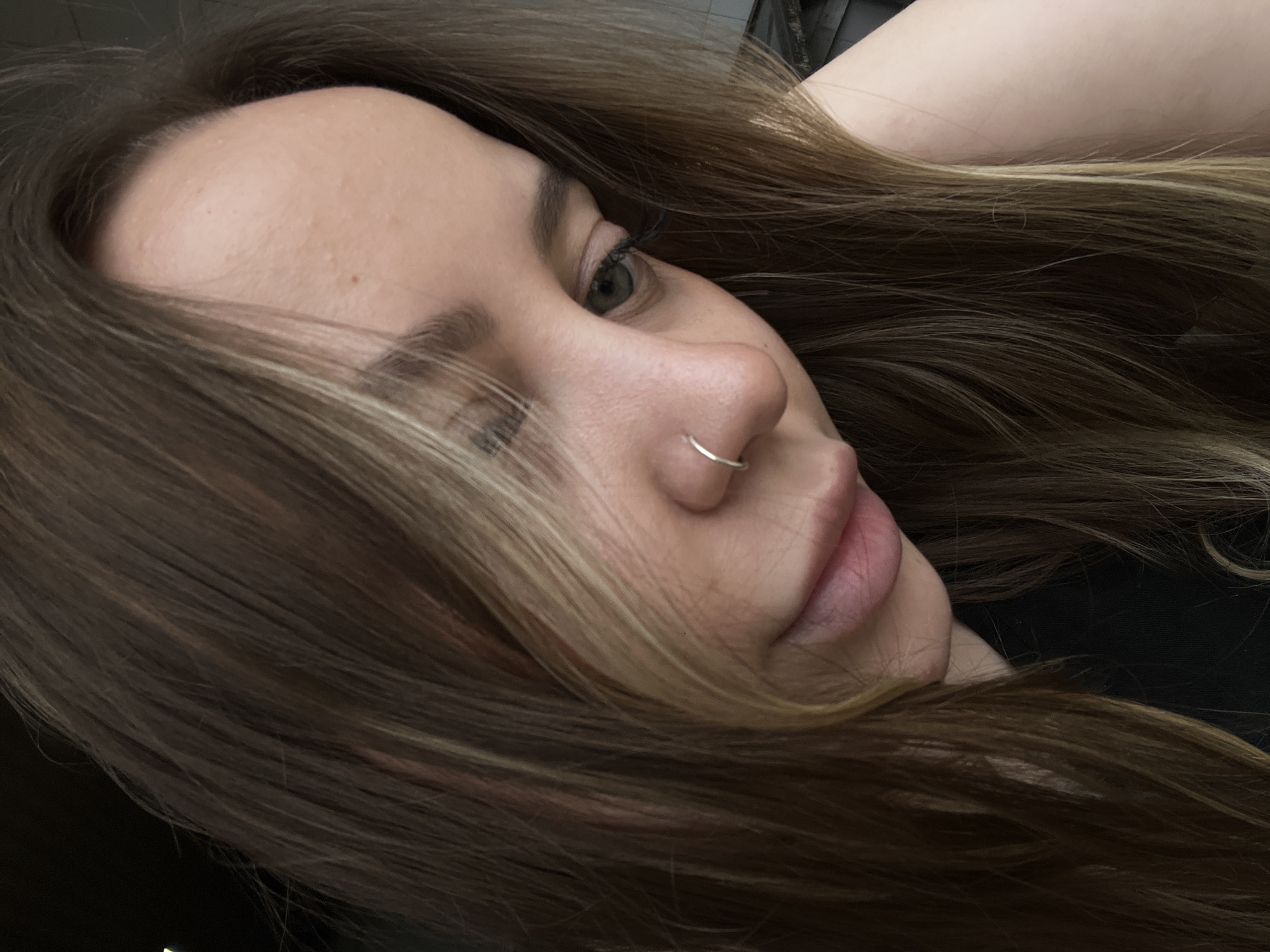 A close-up, tilted selfie of a woman with light brown hair with blonde highlights. She has fair skin, green eyes, and a silver nose ring. Her hair is loose and frames her face. The background is blurred and indistinct, appearing to be a dark color. There is no indication of the location being Novopokrovskaya, Russia.