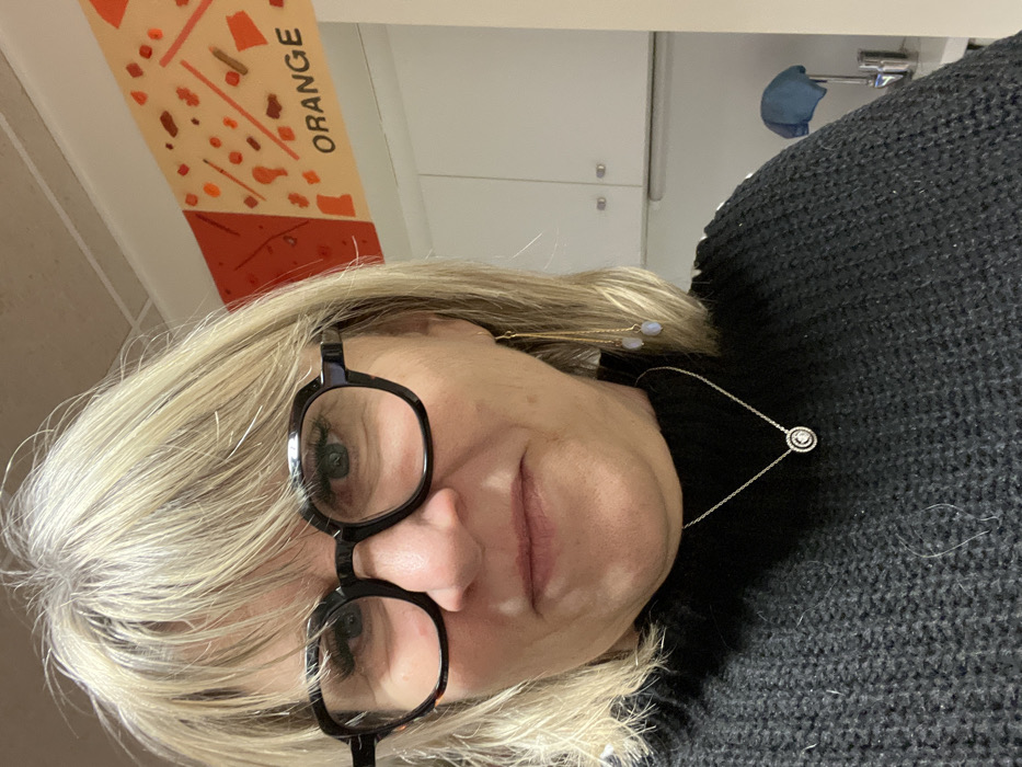 A woman with blonde hair and black-framed glasses is looking directly at the camera. She is wearing a black sweater and a delicate silver necklace. An "ORANGE" sign with abstract shapes is visible in the background, along with a cabinet. She also wears golden earrings.