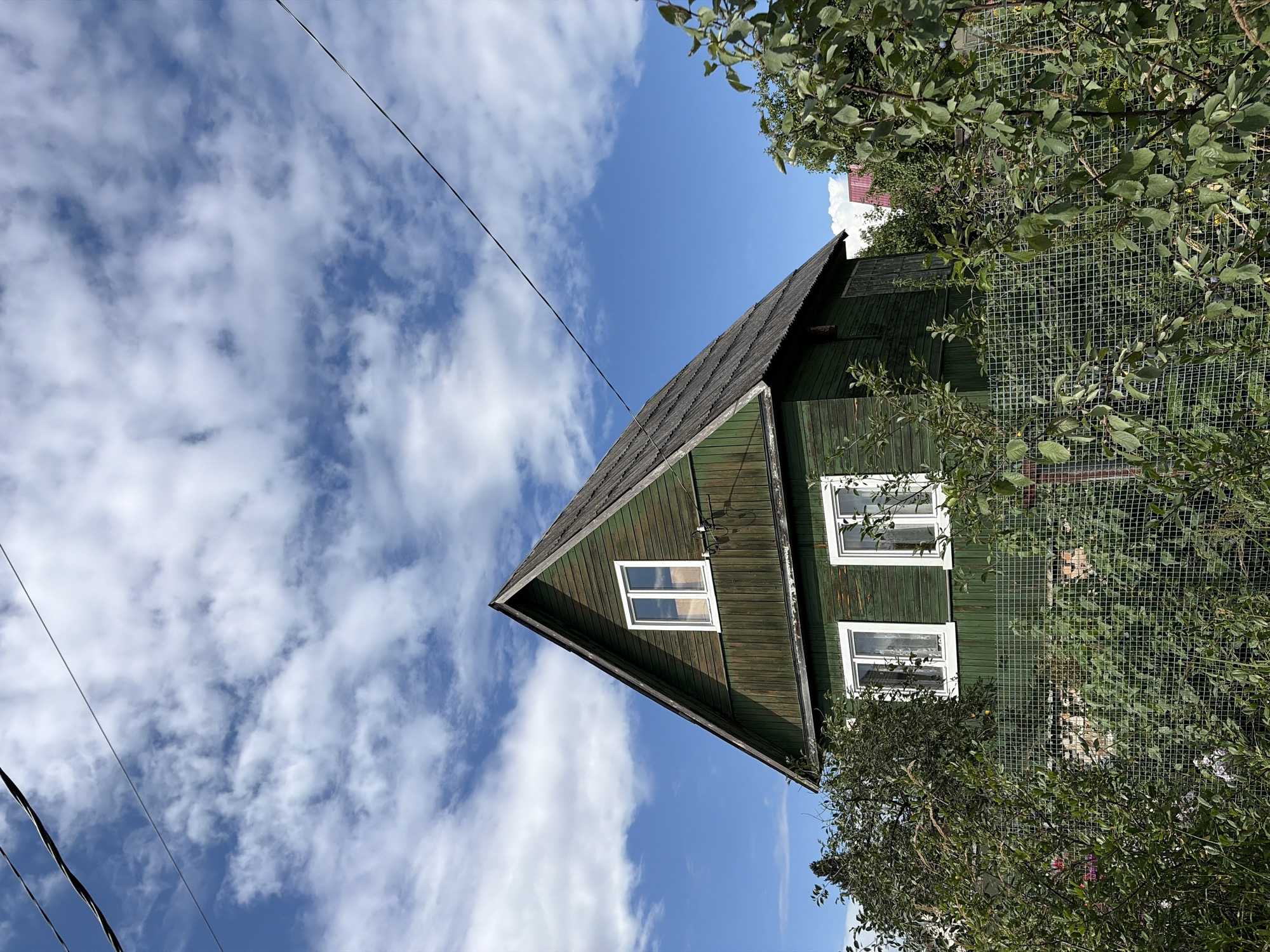 In Нерудник Снт, Russia, a traditional green wooden house with a grey, weathered roof stands under a bright blue sky dotted with fluffy white clouds. Power lines stretch across the upper portion of the frame. The house features a prominent gabled roof and several white-framed windows. Dense green foliage, including trees and bushes, surrounds the house, with a wire mesh fence in the foreground partially obscuring the lower part of the structure and the yard. The overall impression is a rural or suburban setting during daylight.