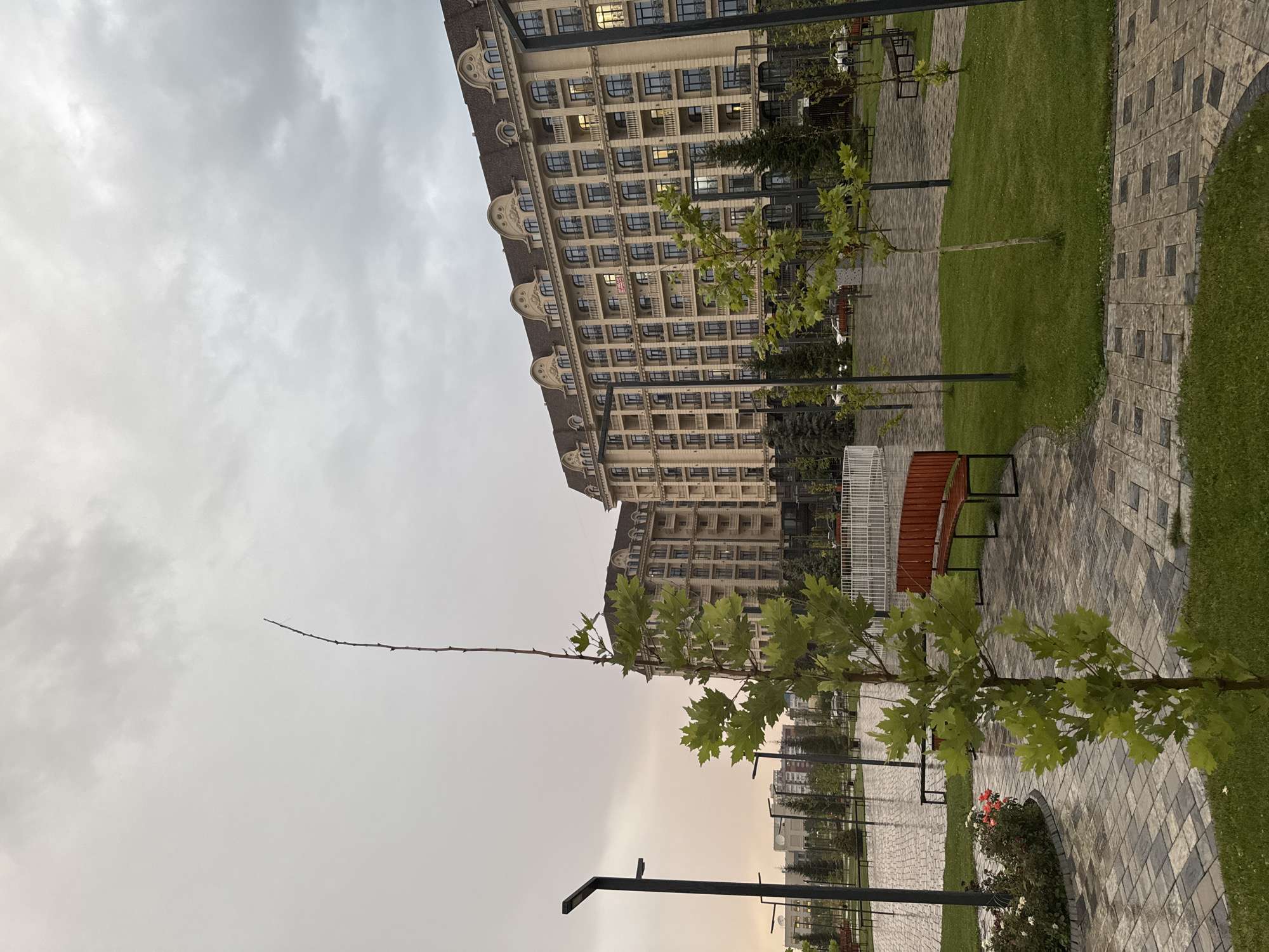 This is an exterior, medium-shot photograph of a residential complex in Bishkek City, Kyrgyzstan, on an overcast day. In the foreground, a young deciduous tree with bright green leaves stands on a patch of lawn. To the left, a circular flower bed contains roses in shades of pink and white. The ground is paved with a mosaic of square stone tiles in varying shades of gray, brown, and black, some of which appear damp. A curved, reddish-brown wooden bench with black metal supports is visible on the right side of the frame. In the mid-ground, more paved pathways, lawns, and trees are present. Streetlights with dark, angular poles are spaced throughout the area. Beyond the park-like setting, two large, multi-story apartment buildings dominate the background. These buildings have light-colored facades, numerous arched windows with dark frames, and ornate decorative elements on their roofs and upper floors. The sky is filled with heavy, gray clouds, suggesting inclement weather. No people are visible in the image. The overall impression is one of a modern, well-maintained urban living space.