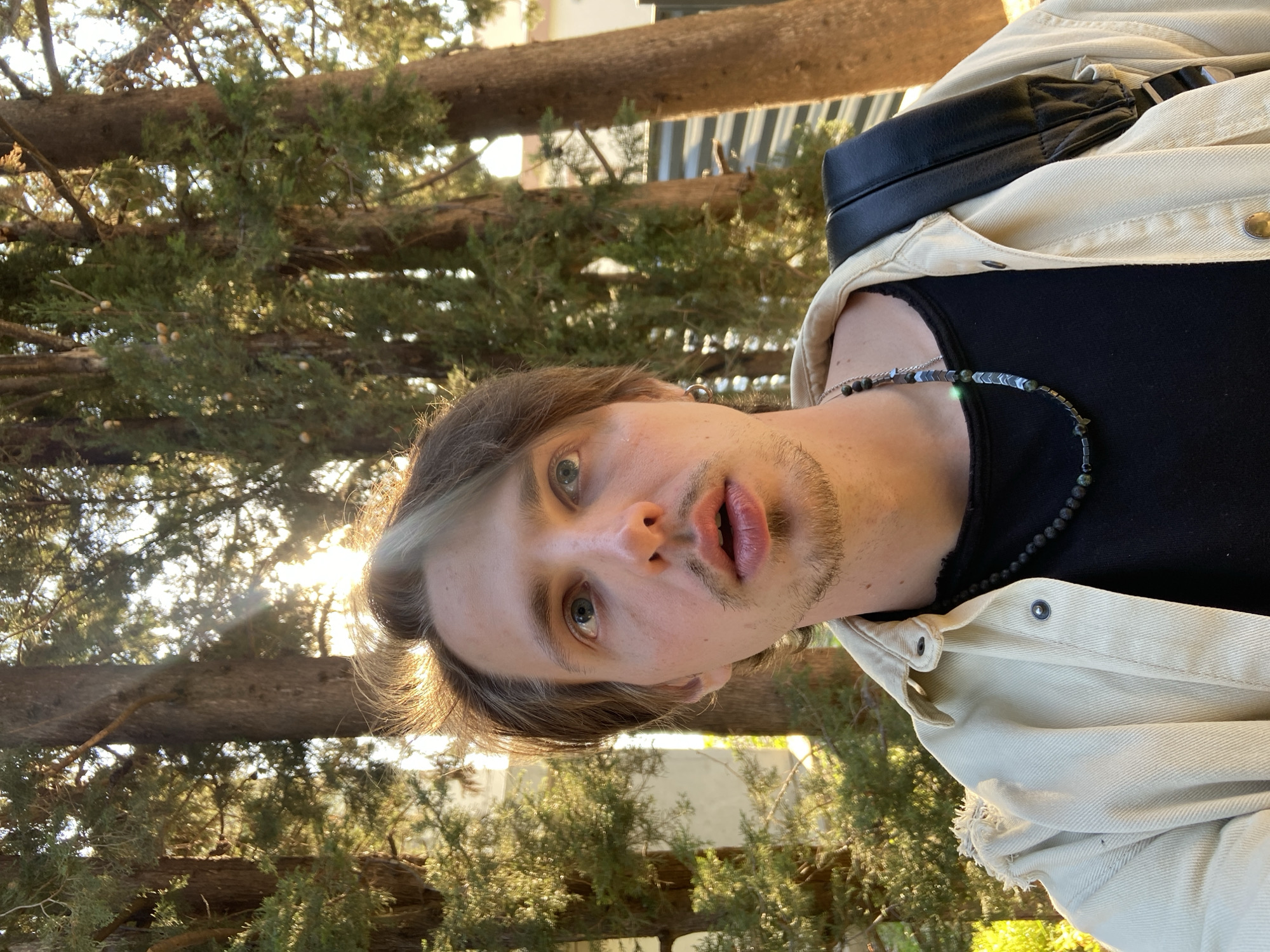 A male with light brown hair with a bleach blonde streak is facing the camera, outdoors. He is wearing a black tank top and a light beige denim jacket, with a black backpack strap visible over his right shoulder. He has a beaded necklace. The background shows tall pine trees and sunlight filtering through the branches, indicating the image was taken in a wooded area. Based on the location information, it can be stated that the photo was taken in a wooded area within the city of Šušanj, Montenegro.
