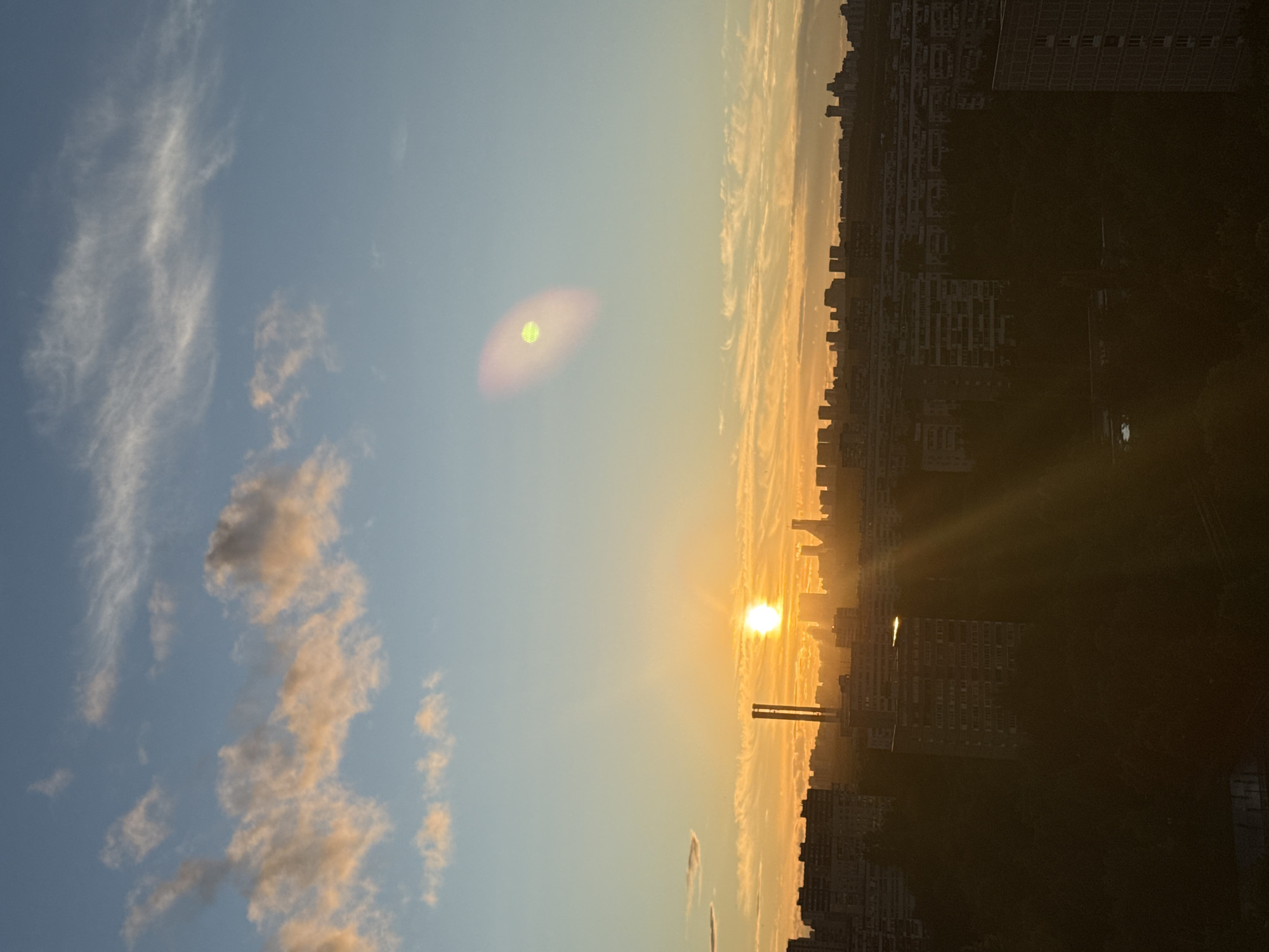 The image captures a sunset over a dense urban landscape in Moscow, Russia. The sun, a bright orb, is positioned low in the sky, casting a warm orange glow that illuminates the clouds and the cityscape. The sky transitions from a pale blue at the top to a vibrant orange near the horizon, with wispy clouds catching the sunlight.
In the mid-ground, silhouetted against the sunset, are rows of apartment buildings and other structures. Two tall, slender industrial chimneys are prominent in the center-left of the frame, likely belonging to a power plant or factory. The buildings vary in height and architectural style, characteristic of a large metropolitan area. Some appear to be older, block-style housing, while others are more modern.
The foreground is dominated by darker silhouettes of trees and more apartment buildings, suggesting the photo was taken from a higher vantage point, possibly a window or balcony of another building. The light rays from the setting sun are visible, fanning out across the scene.
There is a noticeable lens flare in the upper right quadrant of the image, appearing as a large, oval-shaped blur with greenish and purplish hues, indicating the sun's direct or indirect reflection on the camera lens. No people are visible in the image. The overall impression is one of urban sprawl bathed in the dramatic light of a setting sun.