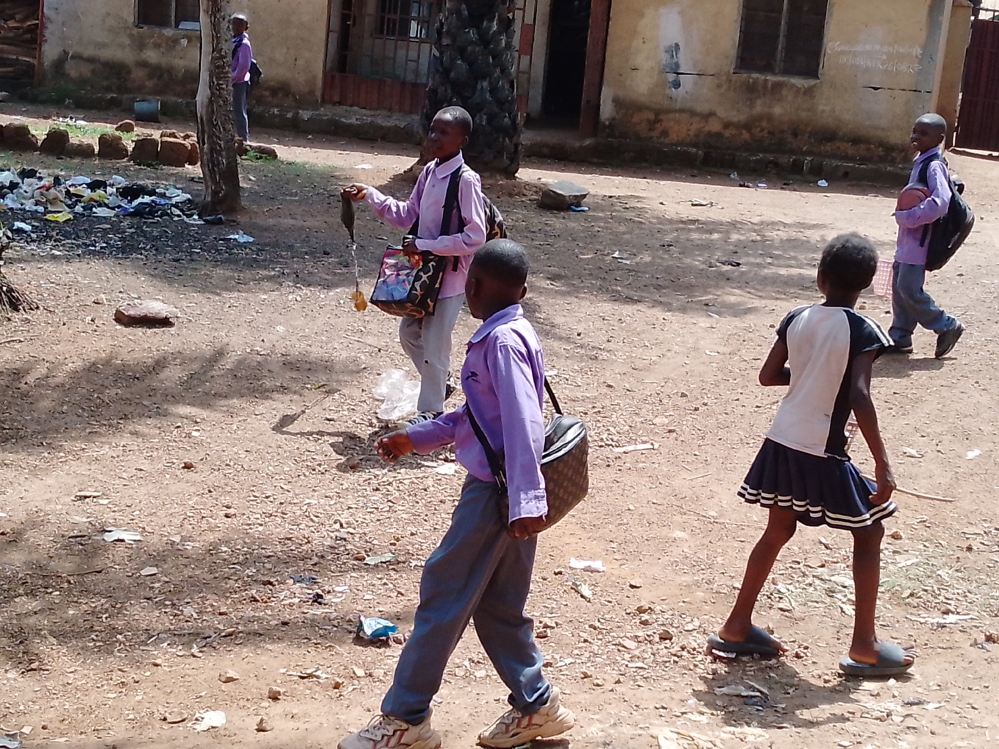 The image captures a scene with multiple children walking along a dirt road in an outdoor setting. The ground is covered with scattered debris and dirt. One child holds an unidentified object attached to a string, and another carries a patterned bag. All children are wearing what appears to be school uniforms. Buildings and vegetation are visible in the background, which also seems cluttered with trash and debris. The location is unspecified, but the scene suggests a developing urban or semi-urban area.