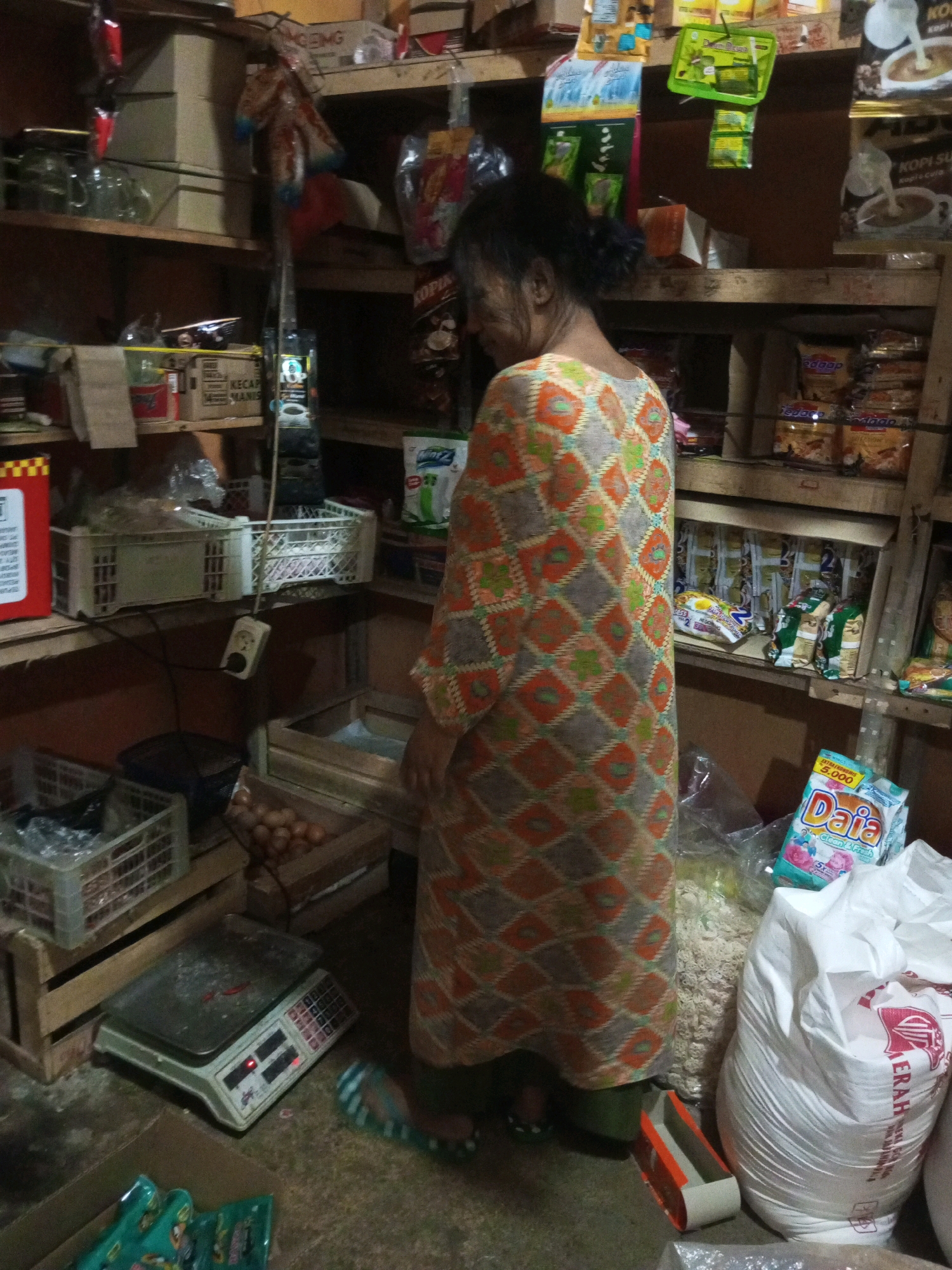 A woman is standing in a dimly lit store filled with shelves stocked with various goods in Andongsili, Indonesia. She is wearing a patterned dress with geometric designs in orange, green, and gray. In the foreground, to the left, is a digital scale, and to the right, a large white sack of what appears to be rice. Shelves behind the woman are laden with packaged food items, including instant noodles and what look like bags of snacks. Crates filled with produce or packaged goods are also visible. The overall environment suggests a small, local convenience store or market stall.
