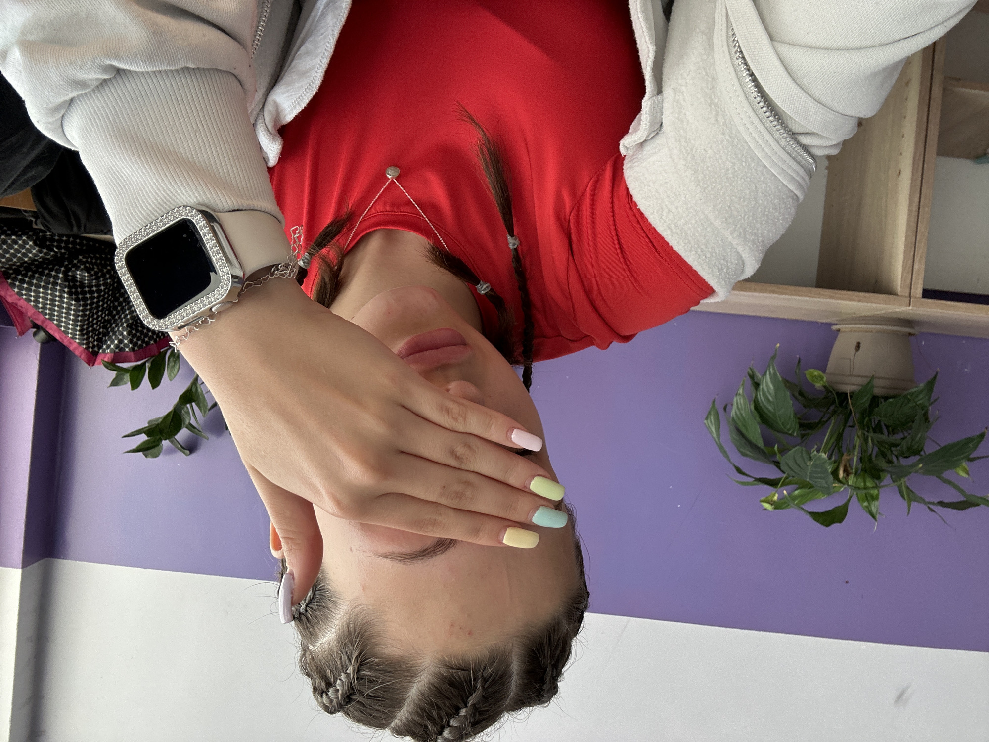 A person is depicted in a sideways orientation, wearing a red t-shirt layered under a light grey hooded sweatshirt. Their left hand is placed over their face, covering their eyes and nose, effectively obscuring their full expression. The fingers display a variety of pastel nail polish colors, including pink, light yellow, light blue, and yellow, with the thumb nail painted white and adorned with a small star. On the left wrist, a smartwatch with a light-colored band is visible, featuring a glittering, possibly diamond-encrusted, bezel. The person's dark hair is styled in multiple small braids or cornrows along the hairline, and a delicate silver-toned necklace with a small circular pendant rests around their neck.
The background consists of a two-toned wall, with the upper section painted a vibrant purple and the lower section in white. In the upper right portion of the frame, a potted plant with lush green leaves, possibly a Peace Lily, can be seen, along with a wooden shelving unit. Another plant or floral arrangement with green leaves and hints of red/pink flowers is partially visible on the far left.
The scene appears to be an indoor setting, likely a room or studio, characterized by the distinct wall colors and decorative plants. The person is posing for a photograph, with the hand-over-face gesture suggesting a playful or shy demeanor, or a deliberate stylistic choice. There is no visible text. The lighting is consistent with an indoor environment during daytime or a well-lit interior. The location context is Sofia, Bulgaria.