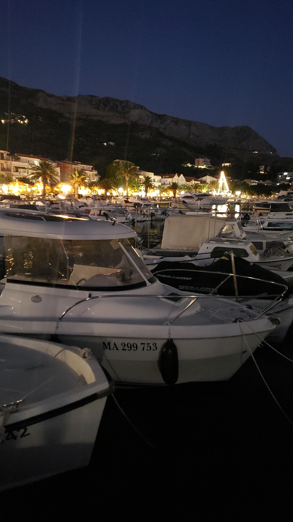 The image captures a nighttime scene at a marina in Općina Tučepi, Croatia. Several boats are moored in the dark water, with a prominent white motorboat in the foreground bearing the registration "MA 299 753." In the background, a town lights up against a dark, mountainous landscape, suggesting a coastal setting. Palm trees line the waterfront, and illuminated buildings and streetlights create a warm glow. A tall, illuminated structure resembling a Christmas tree stands out amongst the buildings. The sky is a deep, dark blue, characteristic of a clear night.