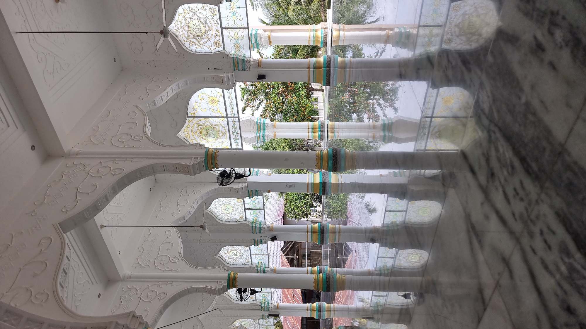 The image depicts the interior of a mosque, likely in Lapang Timu, Indonesia. The foreground is dominated by a highly reflective marble floor, mirroring the architectural elements above. White, fluted columns with decorative bands of turquoise and gold rise from the floor, supporting ornate arches. Intricate white relief carvings adorn the arches and the ceiling. Above the arches, stained-glass windows with floral and geometric patterns, possibly featuring Arabic calligraphy, are visible. Two black oscillating fans are mounted on separate columns. Outside the mosque, through the open arches, trees and the roofs of buildings can be seen, suggesting a daytime setting. The overall impression is one of architectural beauty and tranquility.