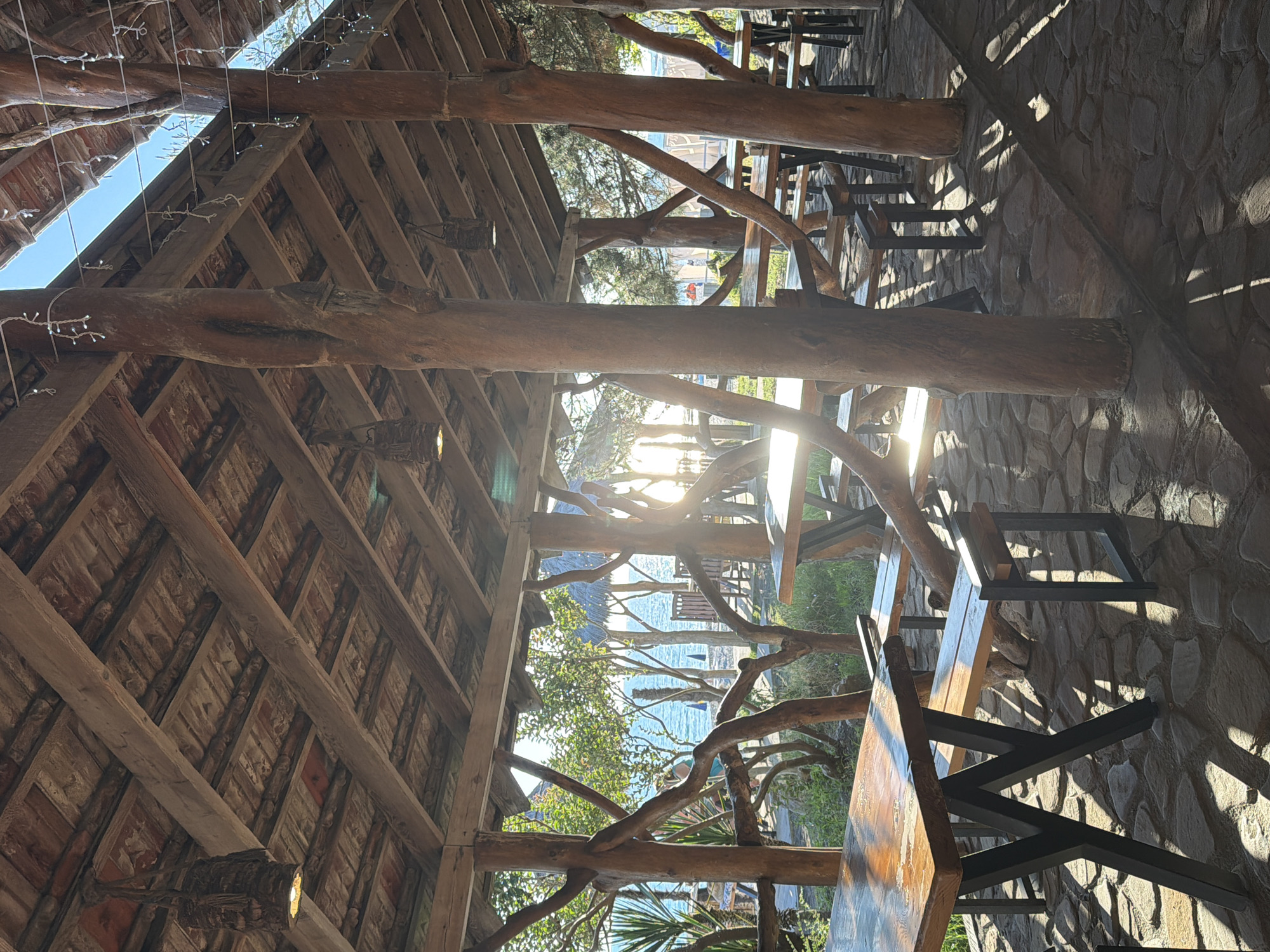 The image depicts an outdoor dining area in Pitsunda, Abkhazia, with a rustic wooden structure providing shade. The roof is made of wooden beams and tiles, with hanging rustic light fixtures. Several wooden tables and benches are arranged on a stone-paved ground. In the background, the blue sea is visible, with trees and vegetation surrounding the area. Sunlight streams through the structure, casting shadows on the ground and tables. No people are clearly visible in the scene.