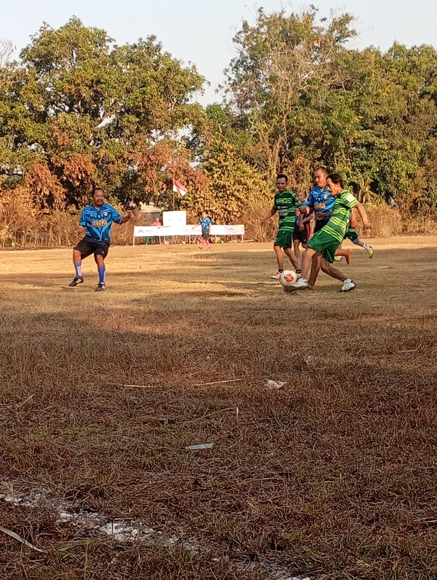 In Kecamatan Haurgeulis, Indonesia, a group of men are playing soccer on a dry, brown grass field. The field is surrounded by trees with dry leaves and some with green leaves, indicating it might be the dry season. In the background, a white banner with text is visible, as well as a red and white flag.
There are at least five men in the frame. One man in a blue and black jersey is standing on the left, looking towards the center of the field. To his right, four men are engaged in playing soccer. Three of them are wearing green and black striped jerseys, and one is wearing a blue and black jersey. The man in the blue and black jersey on the right is kicking the soccer ball. Another man in a green and black striped jersey is positioned to receive the ball, while another man in a similar jersey is to his left. A child in a blue shirt is also visible standing behind the group of players. The men appear to be in their late teens to adult age range. The sunlight suggests it is daytime, likely late afternoon.