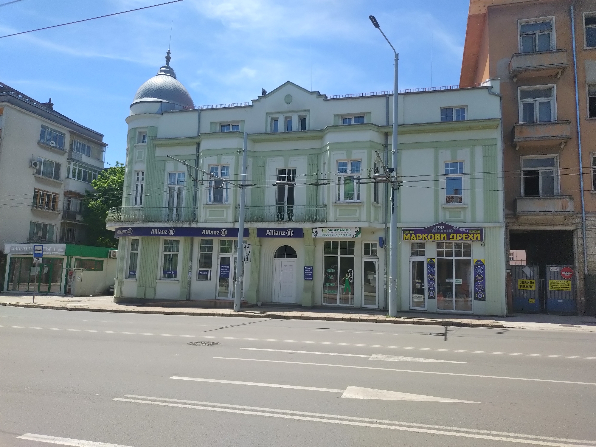 The image depicts a street scene in Stara Zagora, Bulgaria. The buildings are several stories high and feature mixed architectural styles, with classical elements such as columns and a dome contrasting with more modern designs. The building facade facing the street is occupied by several businesses, including an Allianz insurance office, a Salamander shop selling German windows and doors, and a second-hand clothing store. Signs are visible in Bulgarian. The street in front of the buildings is wide and has visible lane markings and crosswalks. There are tram lines above the street. The weather appears clear and sunny. There are no people visible in the image.