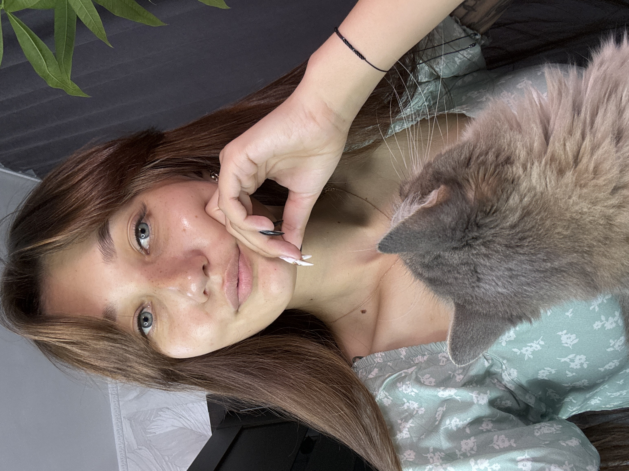 A young woman with long, brown hair and blue eyes is captured in a close-up selfie. She has freckles across her nose and cheeks and is wearing a light green dress with a floral pattern. Her left hand rests on her chin, and she is wearing a black bracelet on her right wrist. A grey cat is partially visible in the foreground, nuzzling against the woman's shoulder. The background is dimly lit, with a dark curtain or wall behind her and a hint of a leafy plant on the right. The setting appears to be indoors, likely a home environment. There is no identifiable information that would suggest this location is in Novokuznetsk, Russia.