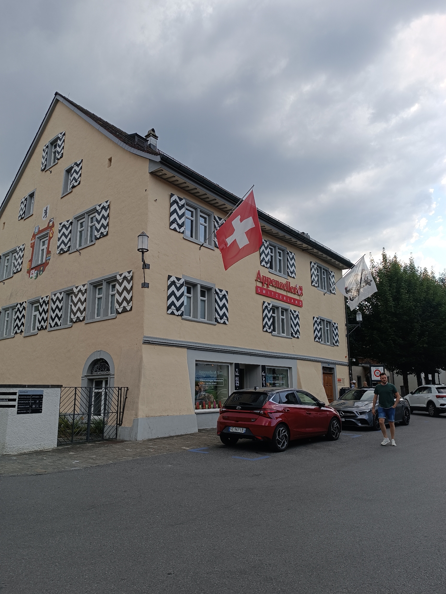 A pale yellow building with a dark, pitched roof stands on a street in Appenzell, Switzerland. The building features an intricate facade with decorative shutters on its windows, painted in a black and white chevron pattern. A large red Swiss flag is prominently displayed on the upper level of the building, alongside another white flag with a crest. The building's ground floor has a storefront with the signage "Appenzelleri & Shop" and displays goods visible through the window. Several cars are parked on the street in front of the building, including a red hatchback, a dark sedan, and a white SUV. A man wearing a green t-shirt and denim shorts walks on the sidewalk, approaching the parked cars. The street is paved with asphalt, and a sidewalk is visible. The sky above is overcast with gray clouds.