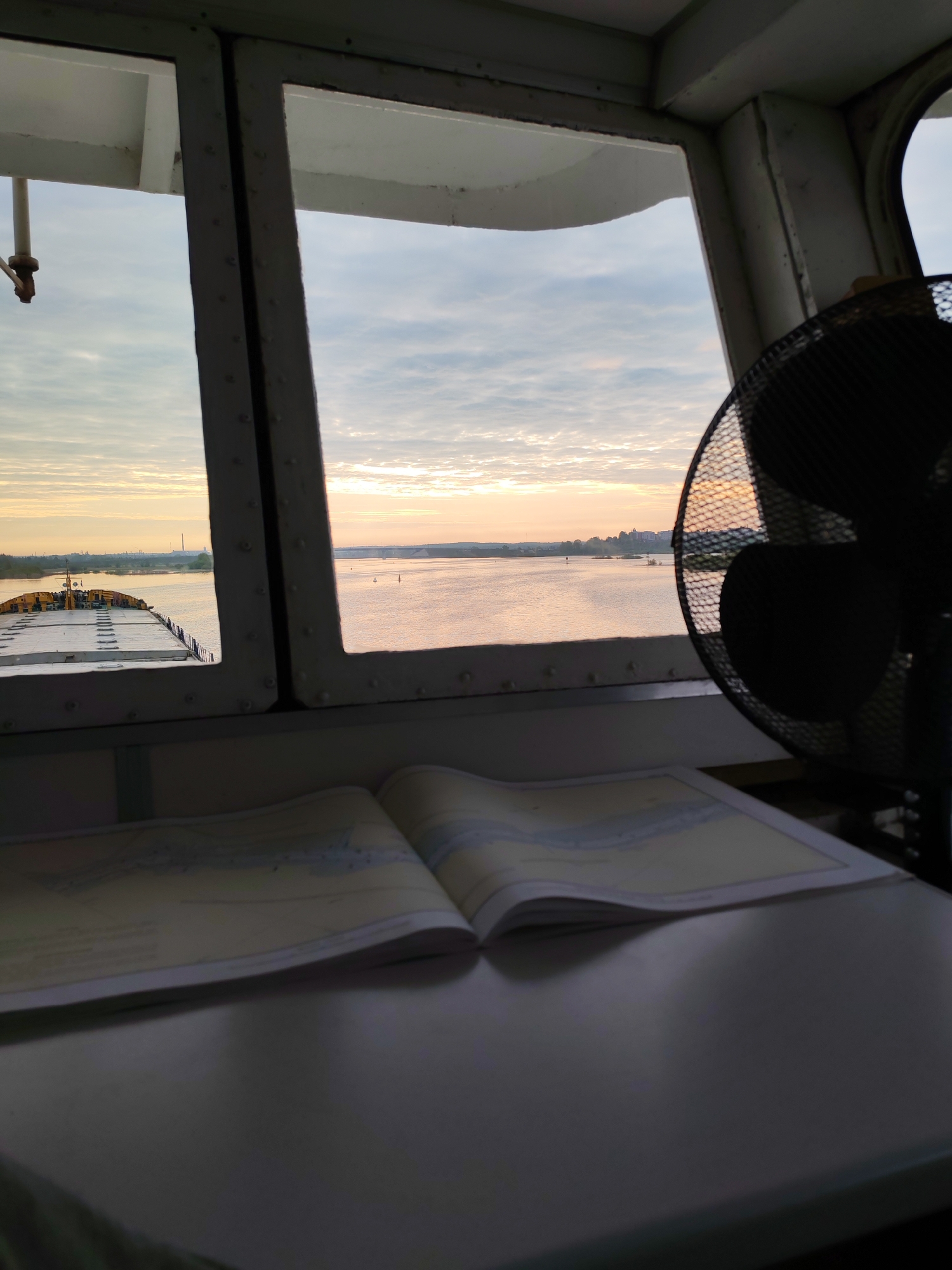 The image shows a view from inside what appears to be a ship's bridge, looking out over a body of water with land visible in the distance. A navigational chart is open on a table in the foreground, and a fan is visible to the right. The sky suggests it is either sunrise or sunset. The location is possibly near Нифантово, Russia.