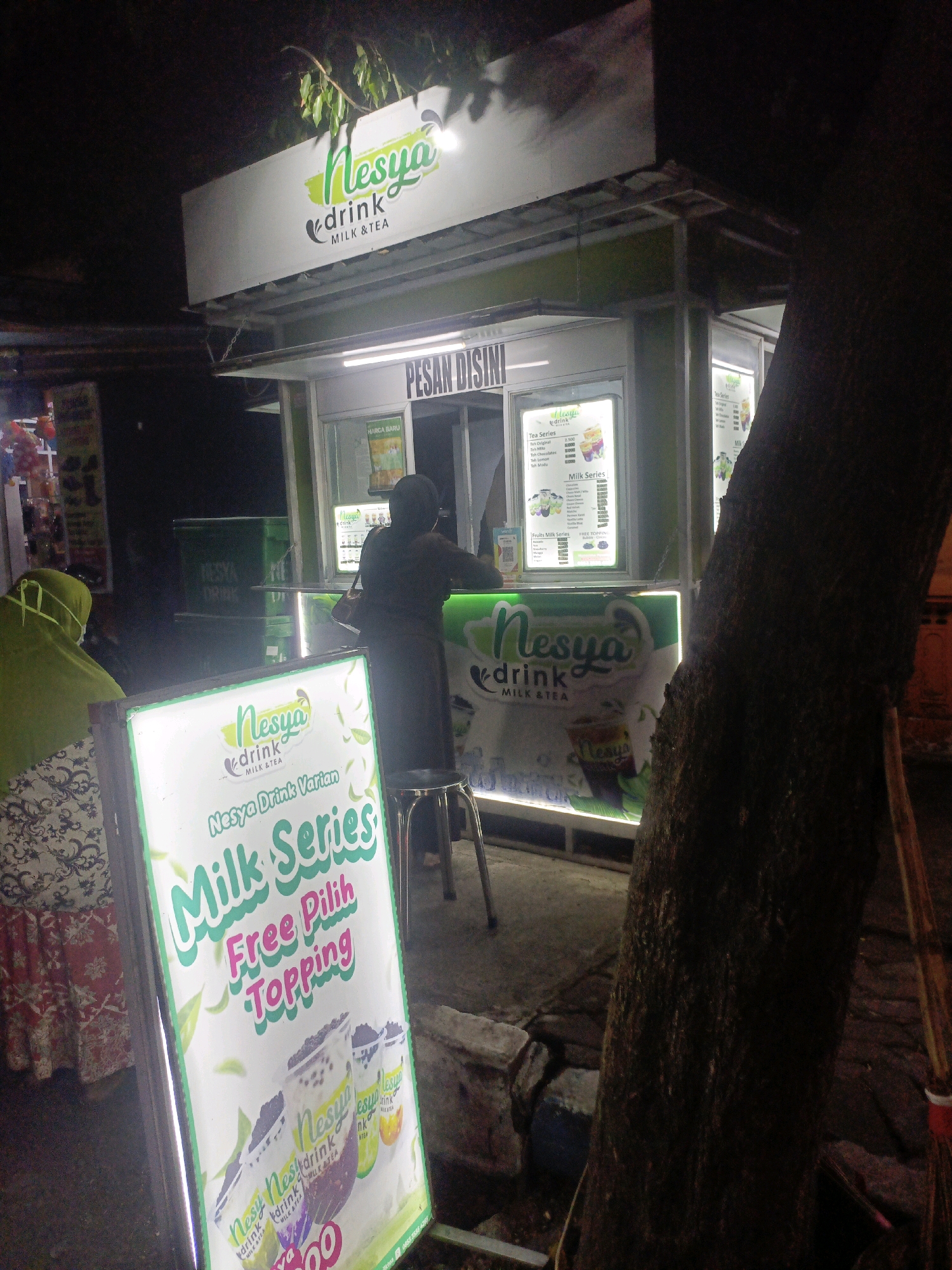The image depicts a street food stall in Probolinggo, Indonesia, at night. The stall, branded "Nesya Drink Milk & Tea," features a brightly lit menu board displaying various drink options. A person wearing a green hijab is seated on the left, facing away from the camera, while another individual, dressed in dark clothing, stands behind the counter and appears to be serving customers. The stall is set up in an outdoor area, with a large tree trunk prominently visible on the right side of the frame, casting shadows. In the background, other stalls and a street lamp are faintly discernible, suggesting a bustling nighttime market environment. The overall scene conveys a casual, local dining experience.