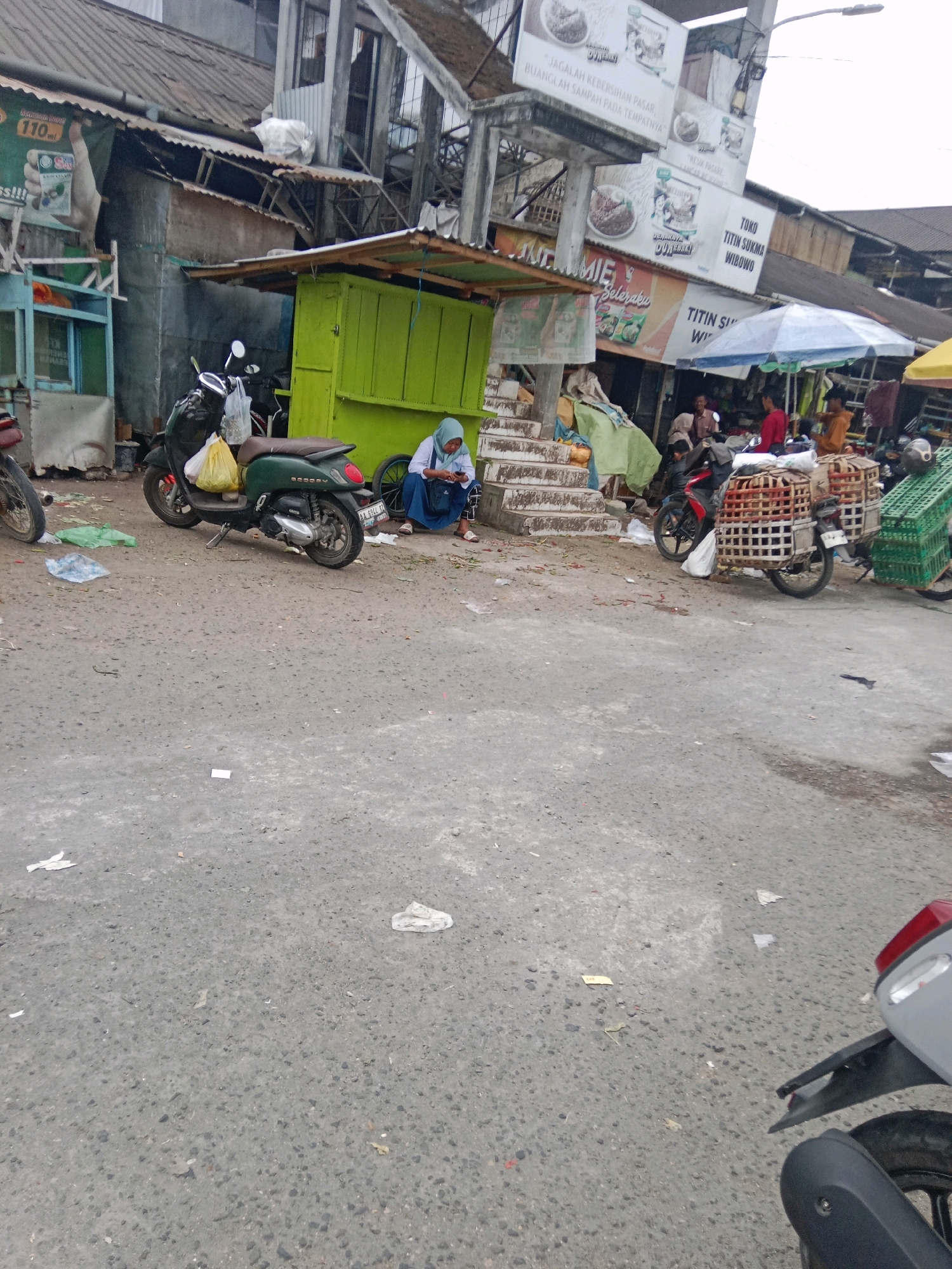 The image depicts a street scene in the city of Kertek, Indonesia, likely a market area, with a focus on the road and the surrounding structures. A green stall with a wooden roof is prominently positioned in the mid-ground. To the left of the stall, a dark green scooter is parked. A woman wearing a light blue hijab and a dark blue skirt is seated on the ground to the right of the stall, looking at her phone. In the background, various buildings and stalls are visible, some with corrugated metal roofs. A large sign with text and images hangs above the market area. Several other motorcycles, one with two large wicker baskets strapped to its back, are also present. The ground is paved with asphalt and littered with debris, including plastic bags and paper. The overall impression is that of a busy, perhaps somewhat disorganized, public space.