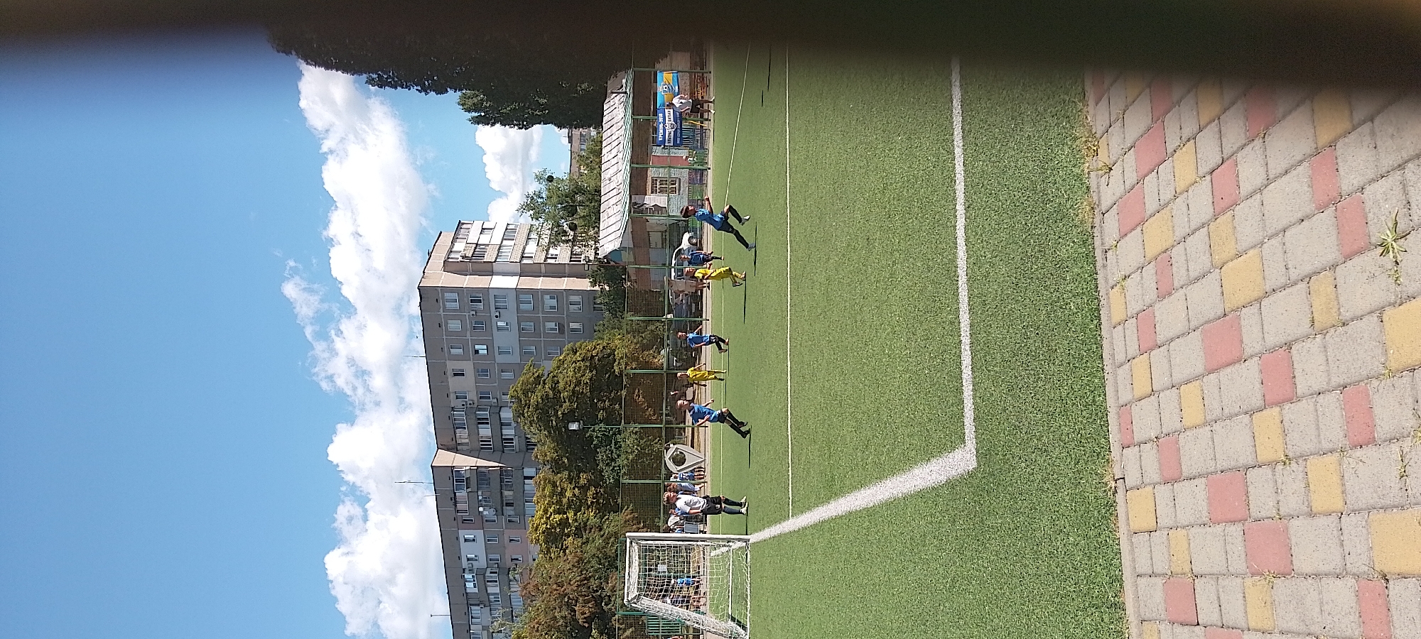 The image depicts a youth football match on a turf field in Cherkasy, Ukraine. The field is marked with white lines, and a goal is visible on the left side of the frame. Several young boys in blue and yellow uniforms are actively playing, running and kicking a ball. In the background, a multi-story residential building and trees are visible. The sky is blue with scattered clouds. The foreground shows a pathway made of interlocking paving stones in red and yellow colors. The overall scene is of a sunny day sporting event for children.