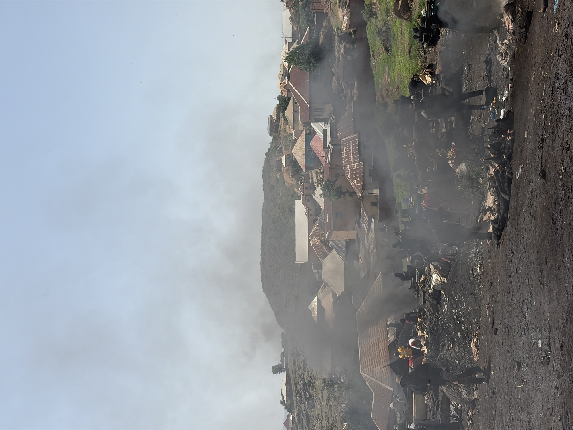 The image depicts a scene of debris and smoke near a hillside settlement in Jos, Nigeria. Smoke obscures the background and some of the immediate foreground. There are visible dwellings or structures with red roofs along the hillside. The foreground is a dirt surface, possibly a road or path, covered with debris. People are present amidst the debris, though obscured by the smoke. The sky is overcast.