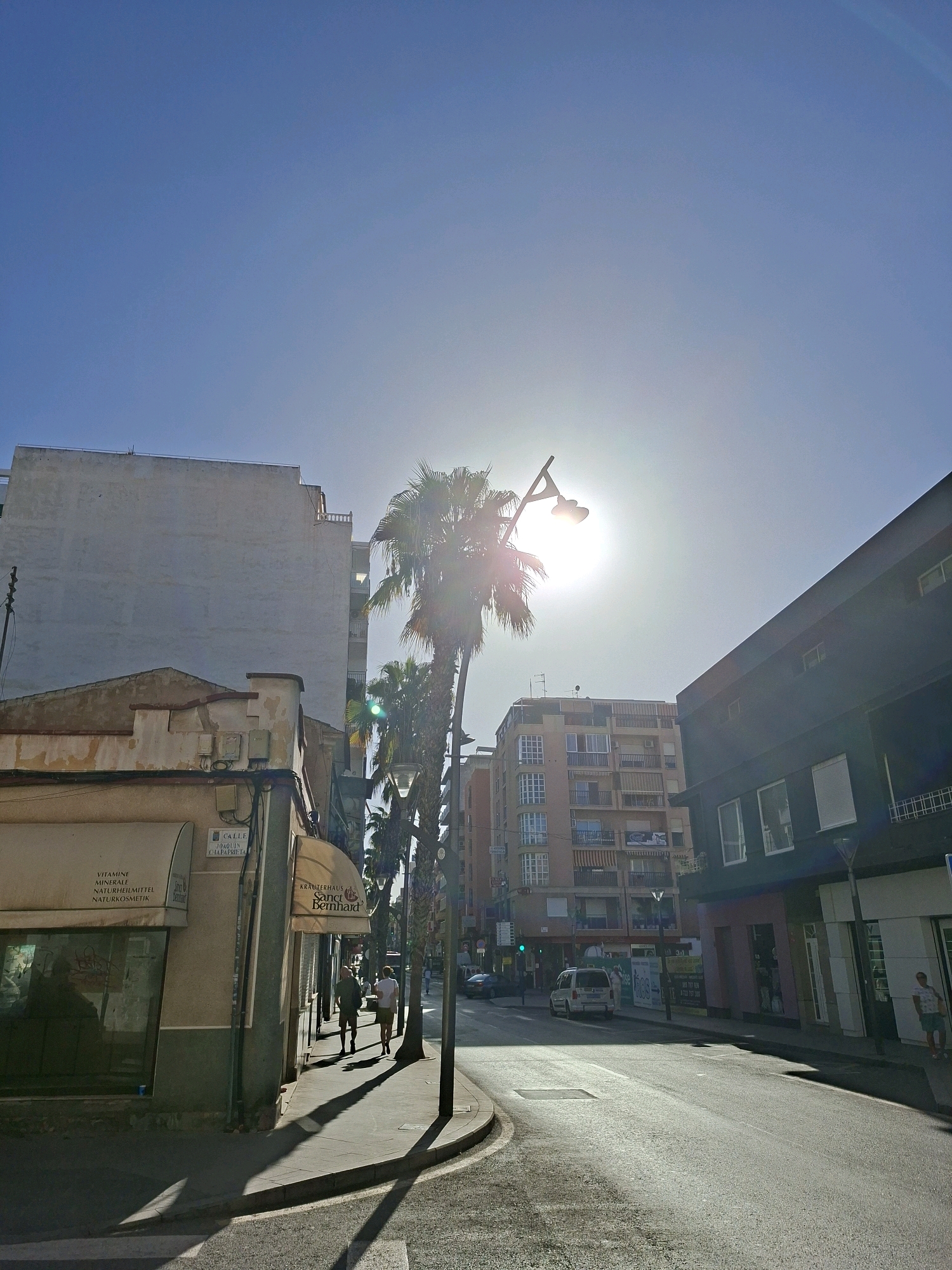 The image shows a street scene in Torrevieja, Spain, under a bright, sunny sky with the sun positioned high and slightly to the right of the center. Tall palm trees line the street, their fronds casting long shadows on the pavement. Buildings of varying heights and architectural styles flank the road. On the left, a light-colored, multi-story building with a blank facade dominates the foreground, adjacent to a smaller, older building with a cream-colored exterior and awnings. A street sign on this building reads "CALLE JOAQUIN CHAPAPRIETA". Further down the street, a tall, tan apartment building with numerous balconies and windows is visible. To the right of this, a dark, modern building with large windows and white blinds stands. The street itself is paved and appears to be a two-way road. Several vehicles, including a white van and a dark car, are parked or driving on the road. Two individuals, a man and a woman, are walking on the sidewalk in the mid-ground on the left side of the street. The overall impression is a typical urban street scene in a sunny climate.