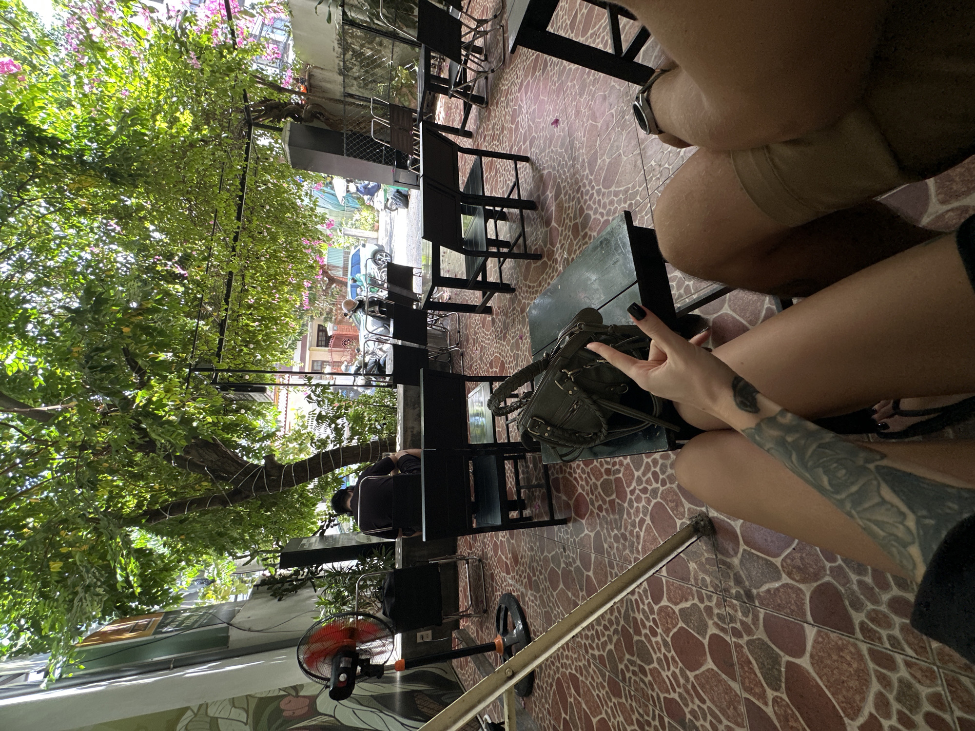 The image captures an outdoor seating area of a cafe in Da Nang, Vietnam, bathed in dappled sunlight filtering through lush green trees and pink flowers. The patio is paved with reddish-brown tiles arranged in a mosaic pattern. Several black metal tables and chairs are arranged throughout the space, some occupied and some empty.
In the foreground, a person's legs and tattooed arm are visible, with black nail polish on their fingernails. They are holding up a hand with two fingers extended in a peace sign. Next to them, another person's legs are visible, wearing khaki shorts and a watch. A dark green handbag with a braided strap rests on a black table in the center of the frame.
Further back, a person in a dark shirt and shorts sits facing away from the camera, seated at a table. To their left, a red and black oscillating fan stands on the tiled floor. In the background, motorbikes and cars are visible on a street, suggesting the cafe is located in a relatively urban setting. The overall atmosphere appears relaxed and casual, characteristic of a cafe setting.