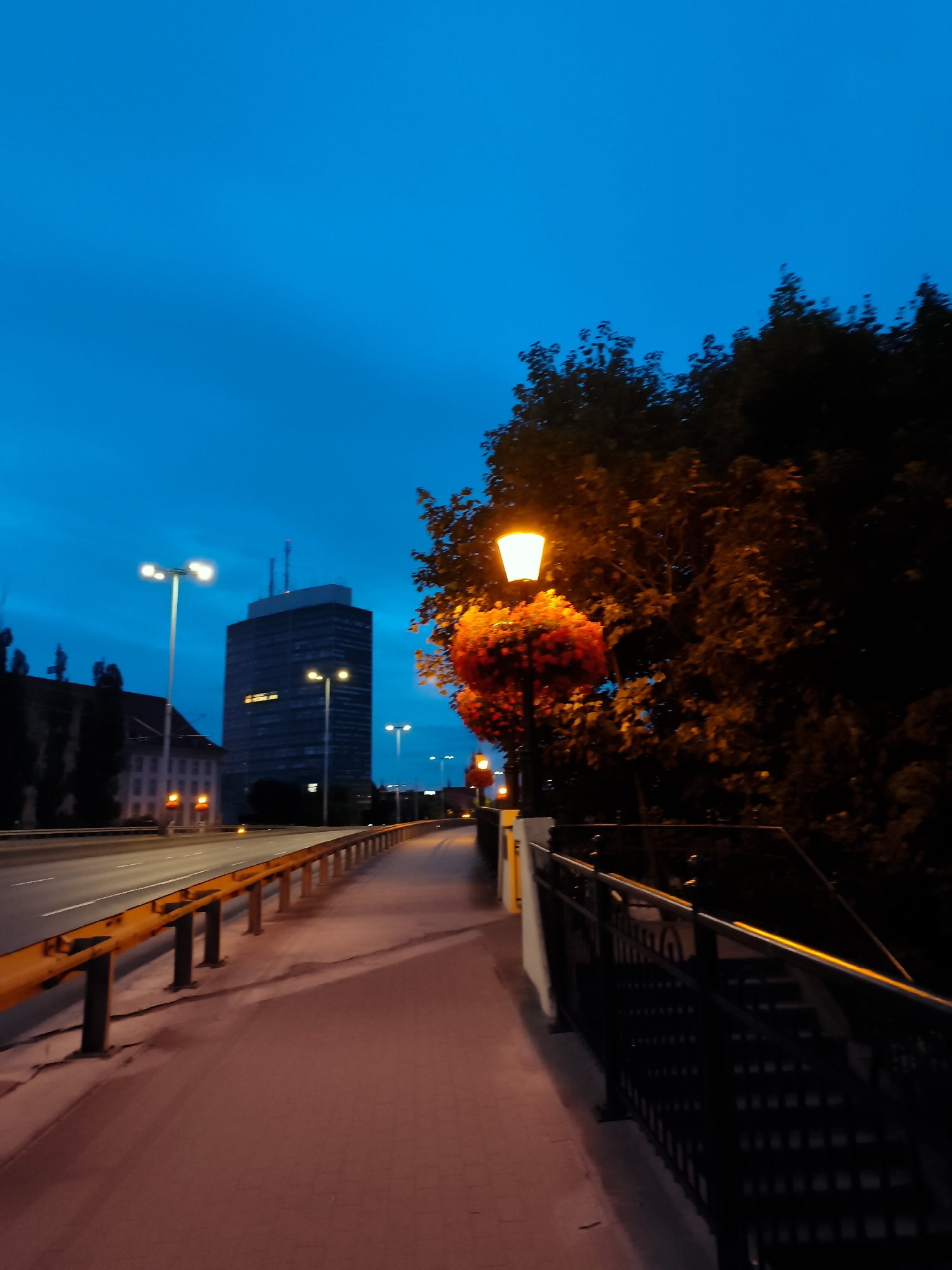 The image depicts a twilight scene on a pedestrian walkway adjacent to a road. In the background, a tall, dark, rectangular building with lit windows stands prominently against a deep blue sky. To the left, there is a lower, lighter-colored building. The walkway is paved with reddish-brown brickwork and lined with a yellow guardrail separating it from the road. A black metal railing runs along the right side of the walkway, leading to a set of stairs. A lamppost adorned with red flowers illuminates the walkway, casting a warm glow. The road itself has visible lane markings and traffic lights. Trees with dark foliage are visible behind the lamppost, adding to the atmospheric ambiance. The overall impression is one of an urban environment at dusk, with the artificial lights beginning to dominate the fading natural light.
