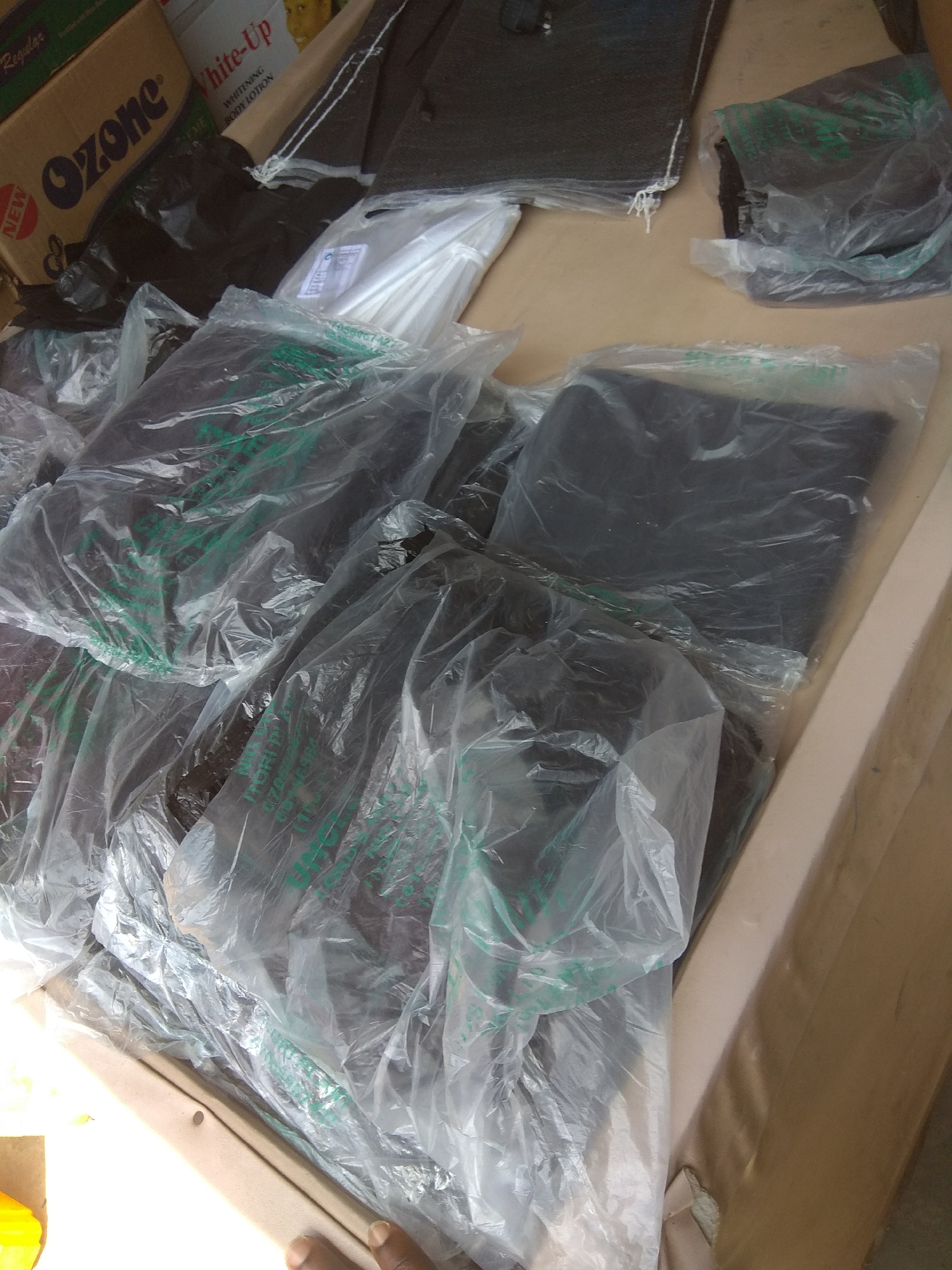 The image depicts a pile of black clothing items, likely garments, neatly folded and individually packaged in clear plastic bags. Some bags have green lettering visible, but the text is not clearly legible. The items are arranged on a flat surface, possibly a table or a shelf, with a cardboard box in the background. The box has visible text that reads "Ozone" in large black letters and "White-Up" in smaller red letters. A portion of a white tub with "WHITENING BODY LOTION" printed on it is also visible on the box. The lighting suggests it is daytime, with natural light illuminating the scene. The overall impression is of stored merchandise, possibly in a shop or warehouse setting. The image appears to have been taken in Jalingo, Nigeria, though no specific location cues are directly evident beyond the provided context. There are no people clearly visible in the image, only a hand in the lower right corner.