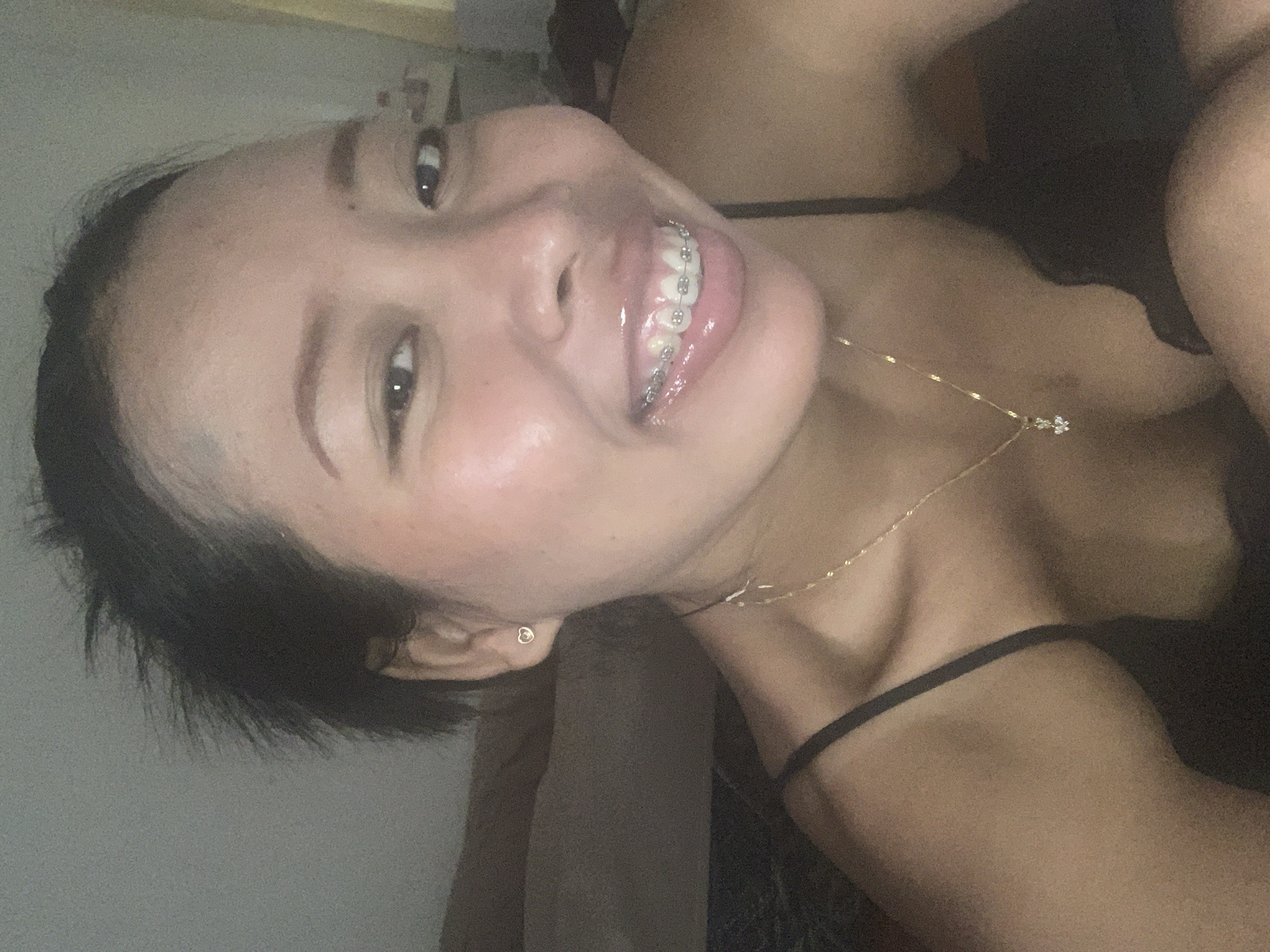 A young woman with short, dark hair is smiling and looking directly at the camera. She is wearing braces on her teeth. She has a small earring in her ear and a delicate gold necklace with a small flower pendant. She appears to be wearing a black tank top. The location is unknown, including the possibility of Denpasar, Indonesia.