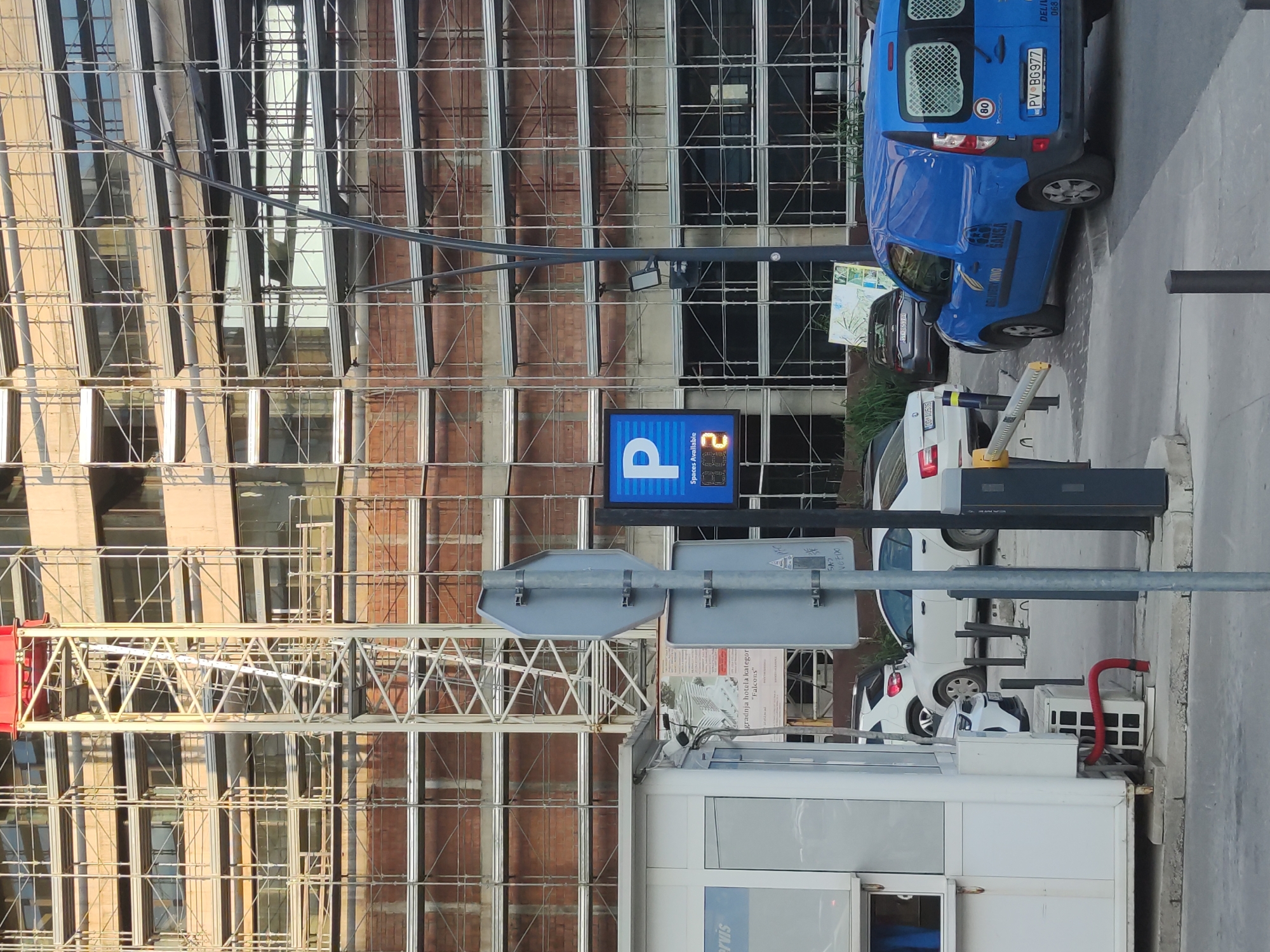 The image captures a street scene in what appears to be a city. In the foreground, a white booth or kiosk is partially visible, adjacent to a parking area. A blue sign with the letter "P" is prominent, indicating parking availability, displaying the number "2". Several cars are parked in the vicinity, including a blue van with license plate "PV BG977". Overhead, a network of scaffolding covers the side of a building, potentially under construction or renovation. Street signs are also present, adding to the urban infrastructure. The overall environment suggests an urban area with ongoing construction or renovation activities.