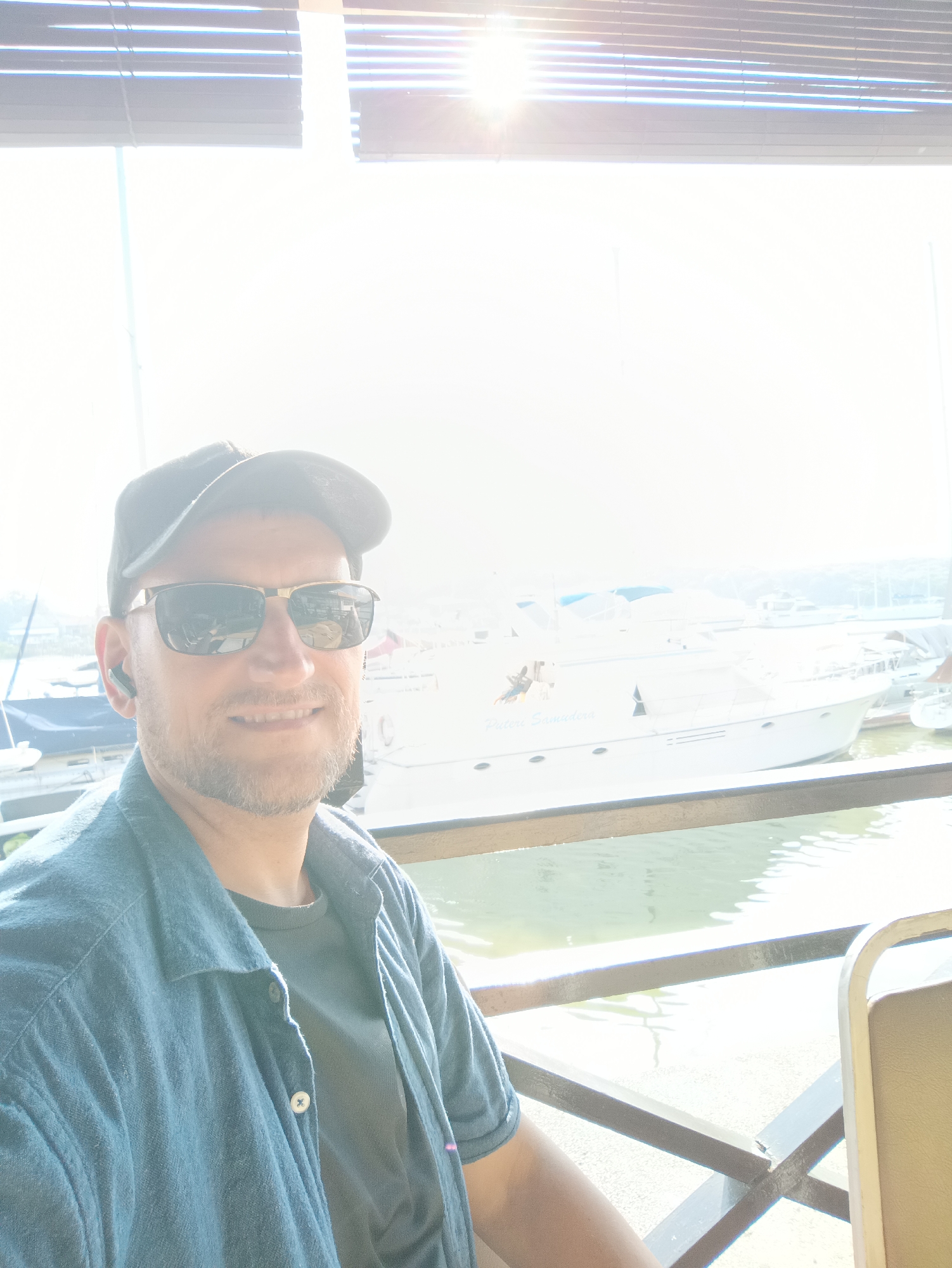 A man wearing a black baseball cap, sunglasses, a blue collared shirt over a dark t-shirt, and an earpiece is taking a selfie. He has a short beard and is smiling. Behind him, a white yacht is docked in a body of water, with other boats and structures visible in the background. The scene appears to be a marina or waterfront area. The lighting suggests it is daytime, with bright sunlight filtering in from the upper right of the frame. This location is identified as Klang City, Malaysia.