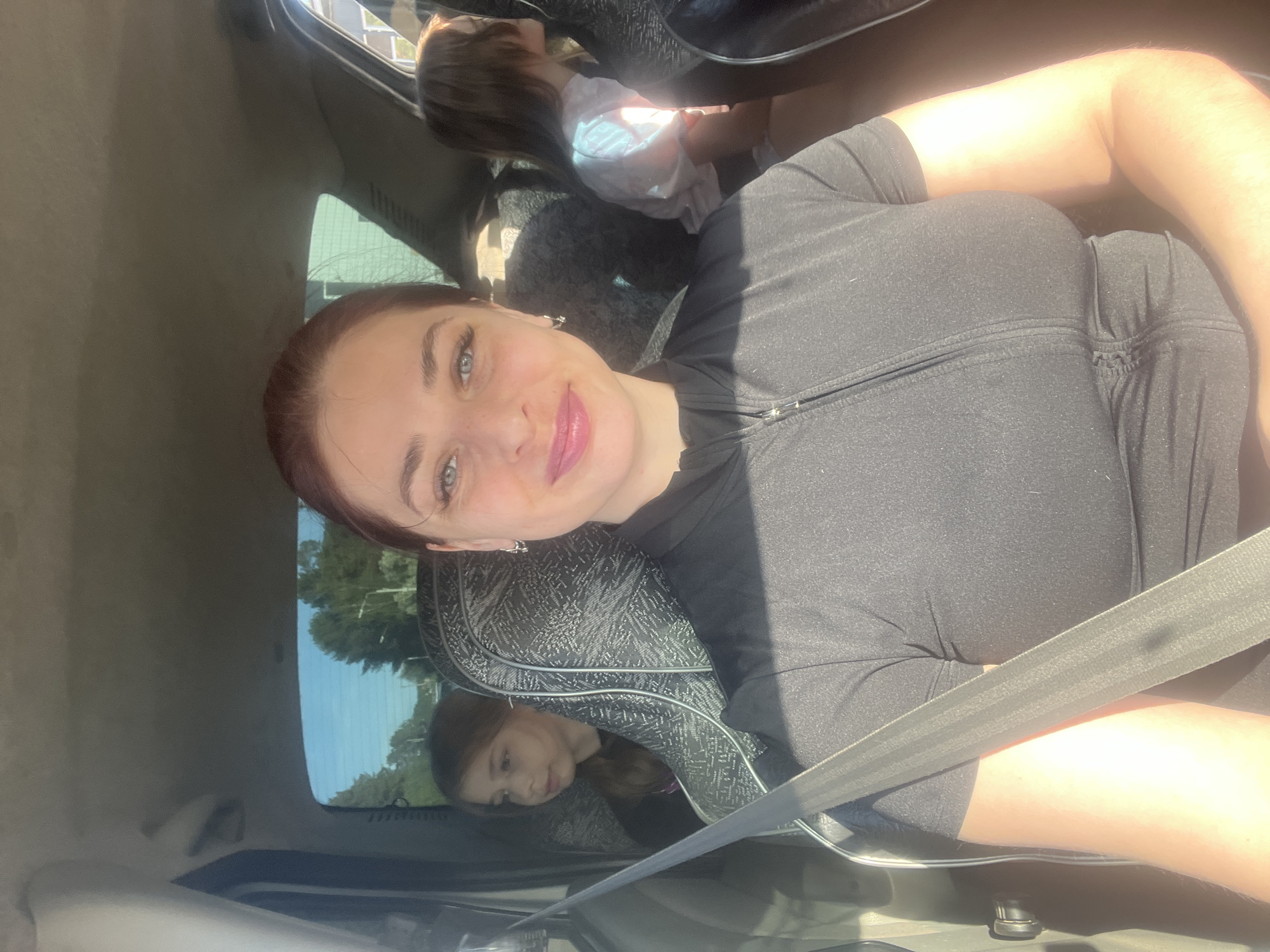 The image shows a woman in the front passenger seat of a car. She is wearing a black top and has a seatbelt fastened across her chest. A child, likely a girl, is visible in the back seat. The car appears to be stationary. The environment outside the car window suggests a suburban or rural area. The location in Chaikovsky, Russia cannot be ascertained from the image alone.