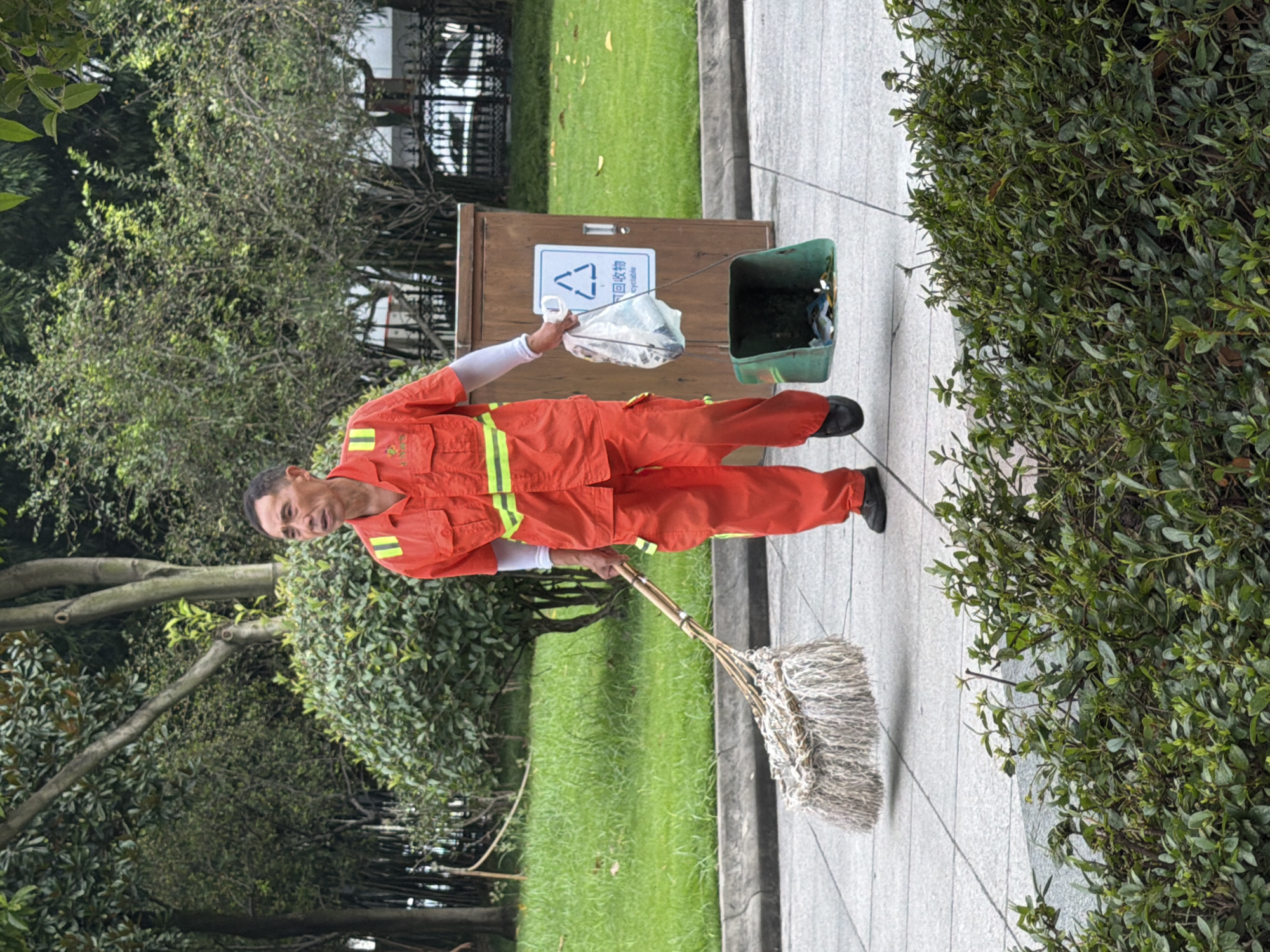 A middle-aged East Asian man, dressed in an orange work uniform with reflective stripes, is walking across a paved pathway in a park. He is holding a large, fibrous broom in his left hand and a white plastic bag filled with refuse in his right hand. A green trash can with some items visible inside is being dragged behind him by a string attached to the plastic bag. To his right, a brown recycling bin with a blue recycling symbol is visible. The environment includes green grass, trees, and bushes. The image appears to be taken in Jiulongpo District, China, based on the provided context.