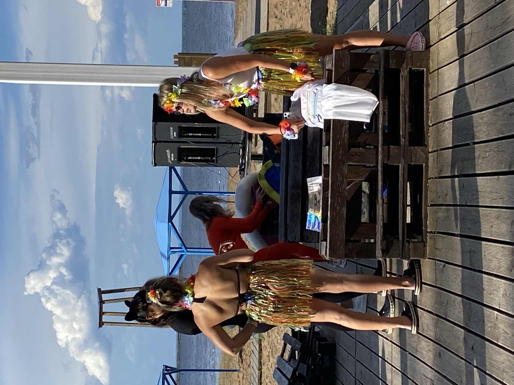 In Chaikovsky, Russia, on a sunny day at a beach, four individuals are present. Two women, wearing leis and grass skirts, appear to be participating in a luau-themed event. One woman, in a white tank top, is reaching into a wooden crate filled with items, possibly preparing for a performance or activity. Another woman, facing away from the camera, is wearing a black bikini top and a colorful grass skirt. She has cat ears on her head. A third person, dressed in a full-body orange jumpsuit, is also interacting with the crate. The fourth individual, partially obscured, is standing behind the person in the bikini. The background features a body of water, a sandy beach, and blue canopies. Two large black speakers are set up on a table. The ground is covered with wooden planks.