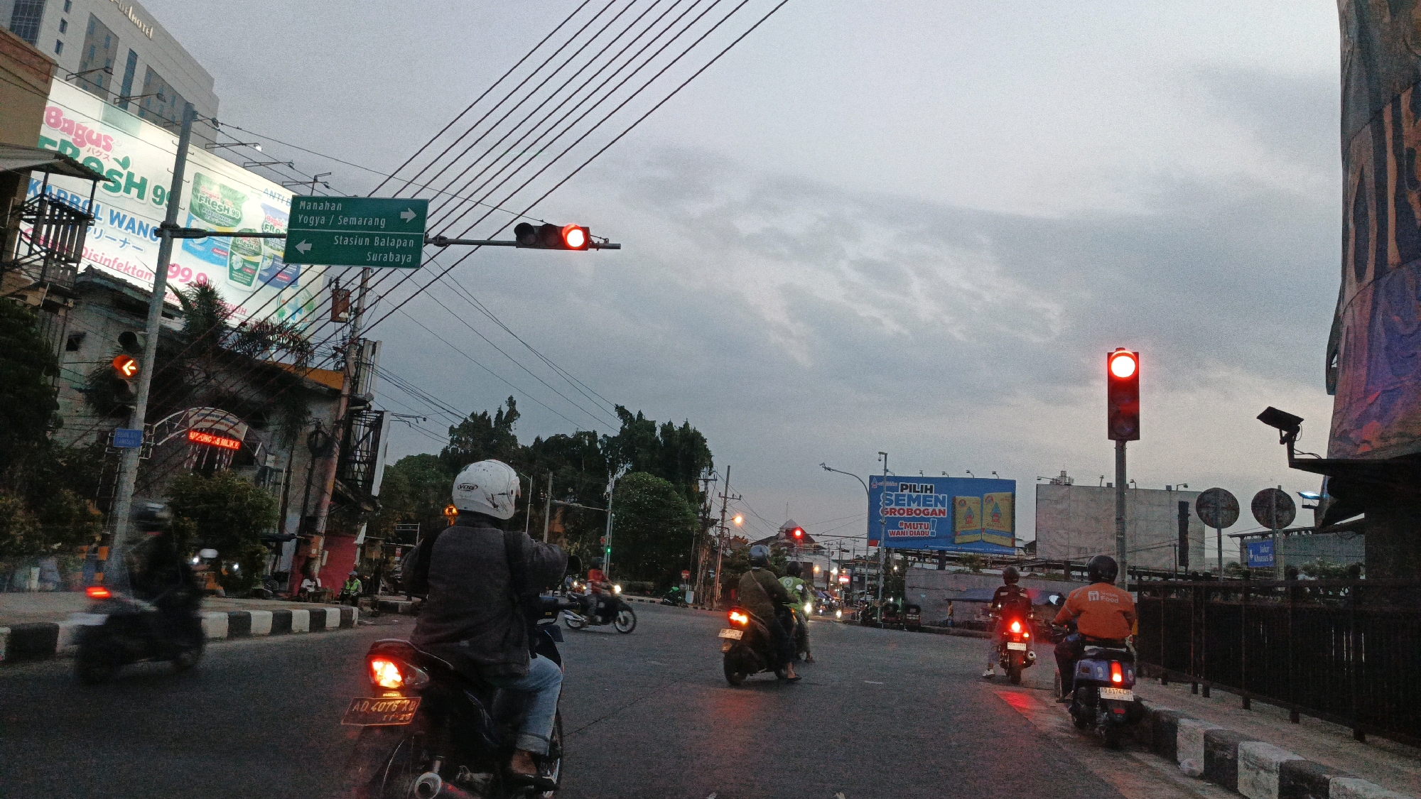 The image depicts a street scene at dusk in Surakarta, Indonesia. A traffic light shows a red signal, causing several motorcyclists to stop. On the left side of the road, a tall building with a sign that reads "Bagus FRESH 99" is visible, along with a billboard. Ahead, a green directional sign indicates "Manahan," "Yogya/Semarang," and "Stasiun Balapan Surabaya." On the right side of the road, another billboard advertising "PILIH SEMEN GROBOGAN" is present. People are riding motorcycles and waiting at the intersection, with trees and electrical wires visible in the background. The sky is overcast.