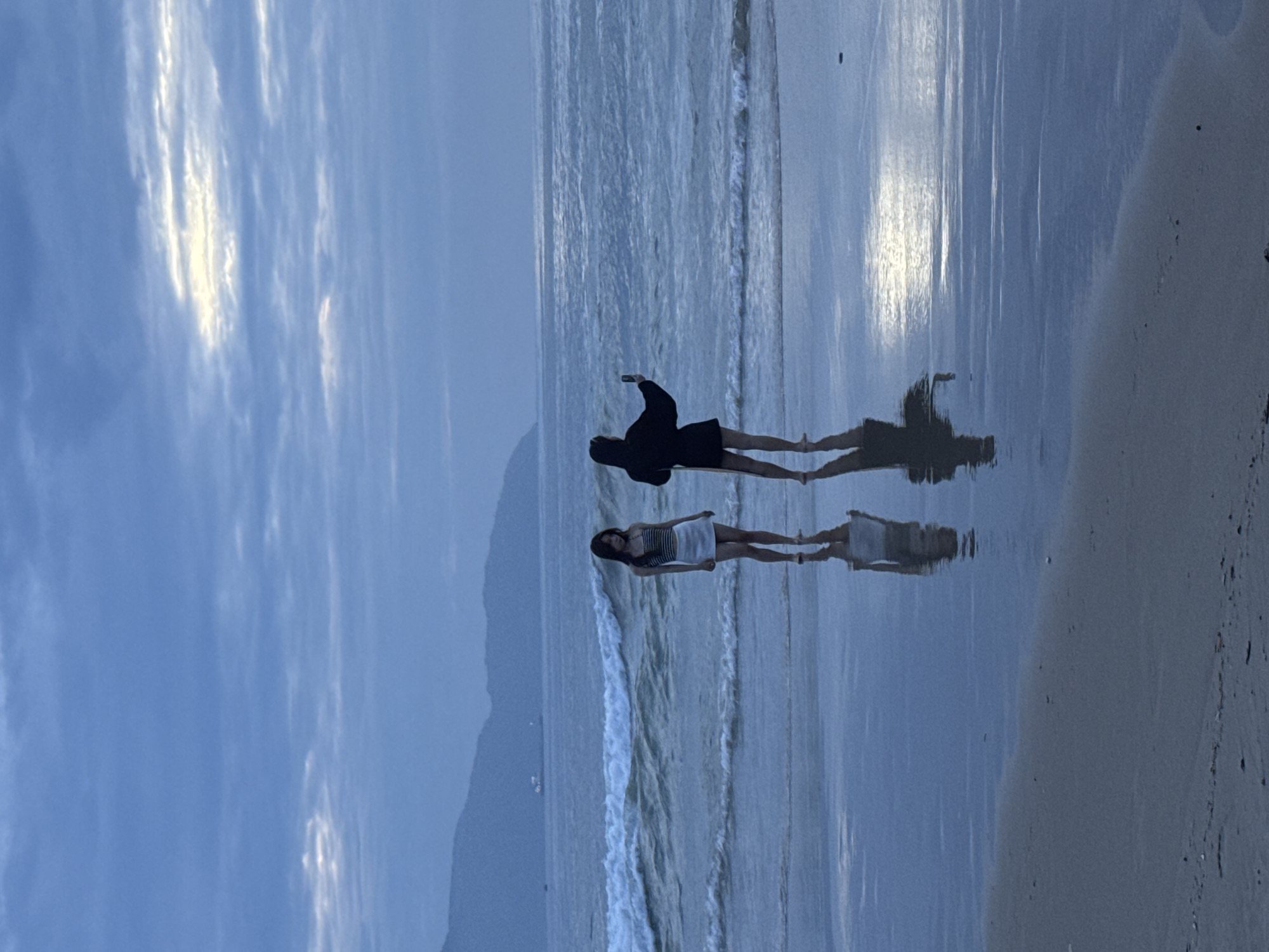 The image shows two people standing on the wet sand of a beach at dawn or dusk. One person wears a dark jacket and appears to be holding a phone. The other wears a light-colored dress. Their reflections are visible on the wet sand. Behind them, waves crash on the shore. Further in the background, a mountain silhouette is visible. The sky is cloudy. The location is My Khe Beach, Đà Nẵng, Vietnam.