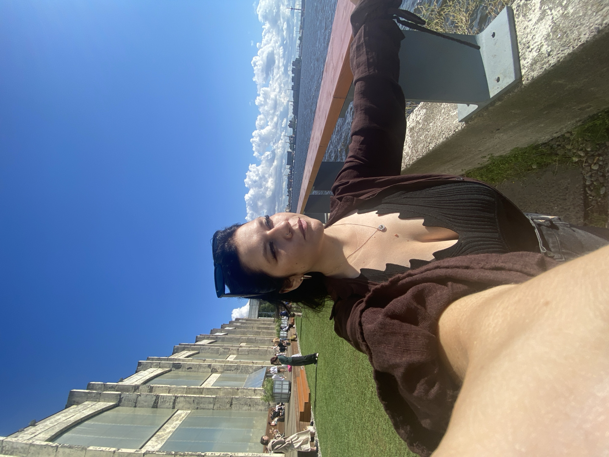 A woman is taking a selfie outdoors on a sunny day. She is standing on a concrete embankment next to a body of water, possibly a river or canal. The embankment has a railing made of wood and metal. Behind her, to the left, is a large, industrial-looking building with tall windows. Further left, on a grassy area, several people are sitting and standing, with some walking. In the background, across the water, is a cityscape with industrial structures and buildings under a blue sky with scattered white clouds. The woman has dark hair and is wearing a dark, ribbed top with a low neckline and a loose, brown open-fronted shirt or jacket over it. She is wearing sunglasses perched on her head. Her right arm is extended forward, holding a phone to take the photo.