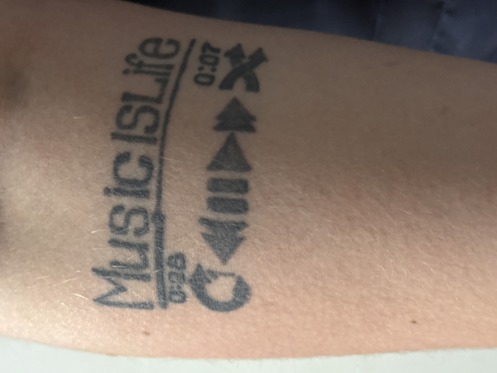 A person's forearm with a black ink tattoo is visible. The tattoo features the text "Music IS Life" with a horizontal line underneath. Below this line, the text "0:06" is present. Further down, a series of symbols are tattooed, resembling audio player controls: a looped arrow, a backward arrow, three horizontal lines, an upward arrow, and crossed arrows, next to “0:07”. No location or person can be identified.