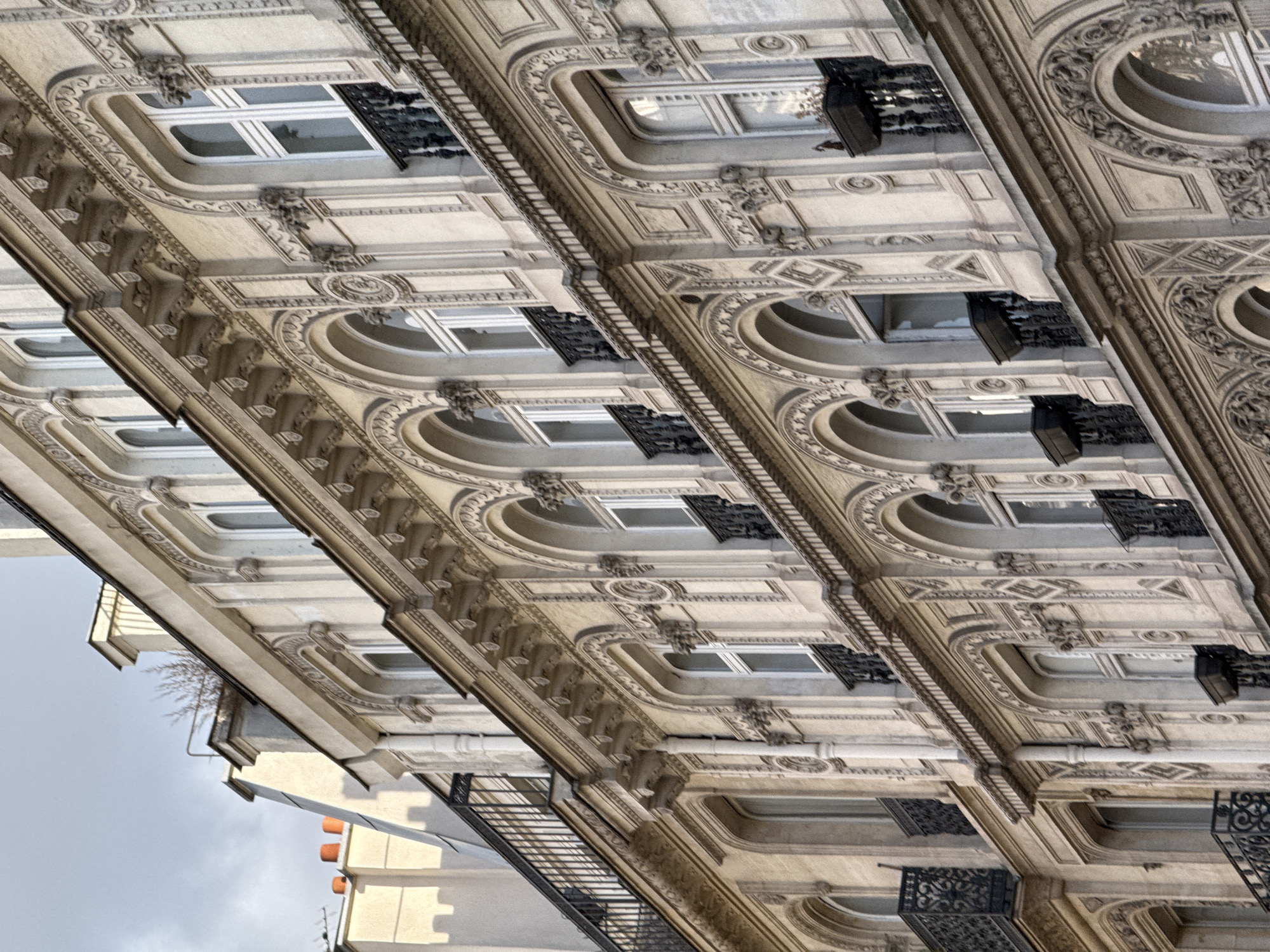 This image captures an artistic, rotated perspective of an ornate building facade, characteristic of Parisian architecture, likely a Haussmannian-style edifice. The photograph, taken from a low angle looking upward and then rotated clockwise, presents the multi-story structure horizontally, emphasizing the repetitive grandeur of its design.
The facade is constructed from light-colored stone or stucco, richly adorned with intricate decorative elements. Each window bay features detailed moldings, curved cornices, and balustrades. Below many windows are small, dark wrought iron balconies or railings with elaborate scrollwork, while some windows are set into deeper, arched recesses. Between window levels, ornate carvings, including classical motifs and figures resembling grotesques, add to the building's lavish aesthetic. A few dark, larger wrought iron structures, possibly parts of fire escapes or more substantial balconies, are also visible.
The scene appears to be set during the daytime under an overcast sky, which provides soft, diffused lighting, highlighting the architectural textures without harsh shadows. In the periphery, parts of a simpler, lighter-colored building and a metal staircase or fire escape can be seen, along with a small, leafless tree or bush growing on a lower building section. No specific activities are occurring, and no text is visible anywhere in the image. The overall impression is one of historical elegance and detailed urban craftsmanship in Paris, France.