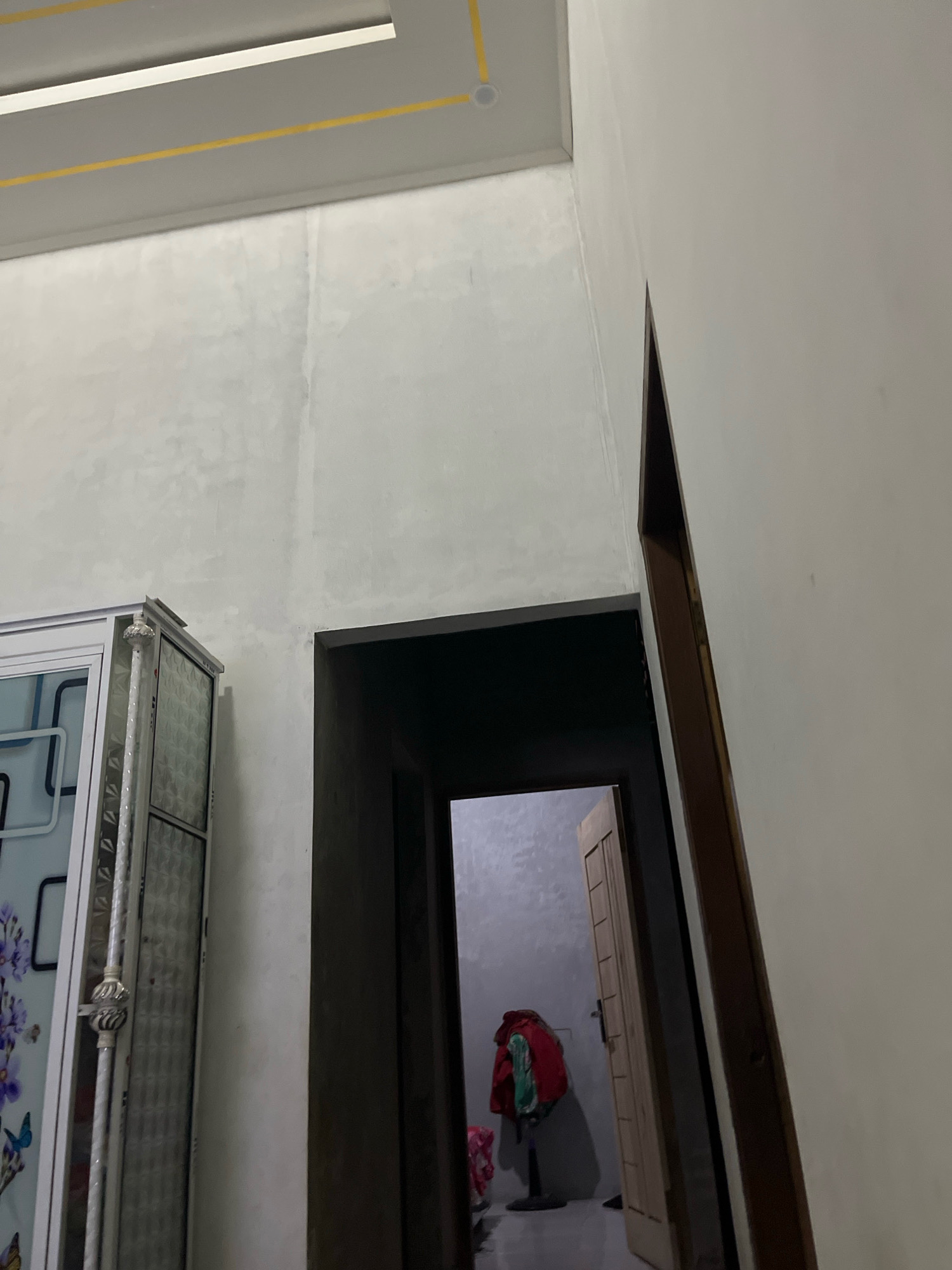 The image depicts an interior scene, possibly a residential hallway or room in Kebumen, Indonesia. In the foreground left, a portion of a white wardrobe with decorative floral and butterfly motifs is visible. To the right of the wardrobe, a dark doorway leads into another room. Beyond the doorway, a wooden door is ajar, revealing a glimpse of the space within. A white fan stands in the adjacent room, with several pieces of clothing, including a red jacket, draped over it. The walls are painted a neutral, light color, and a yellow accent line runs along the ceiling. The lighting appears to be artificial, casting shadows within the space.