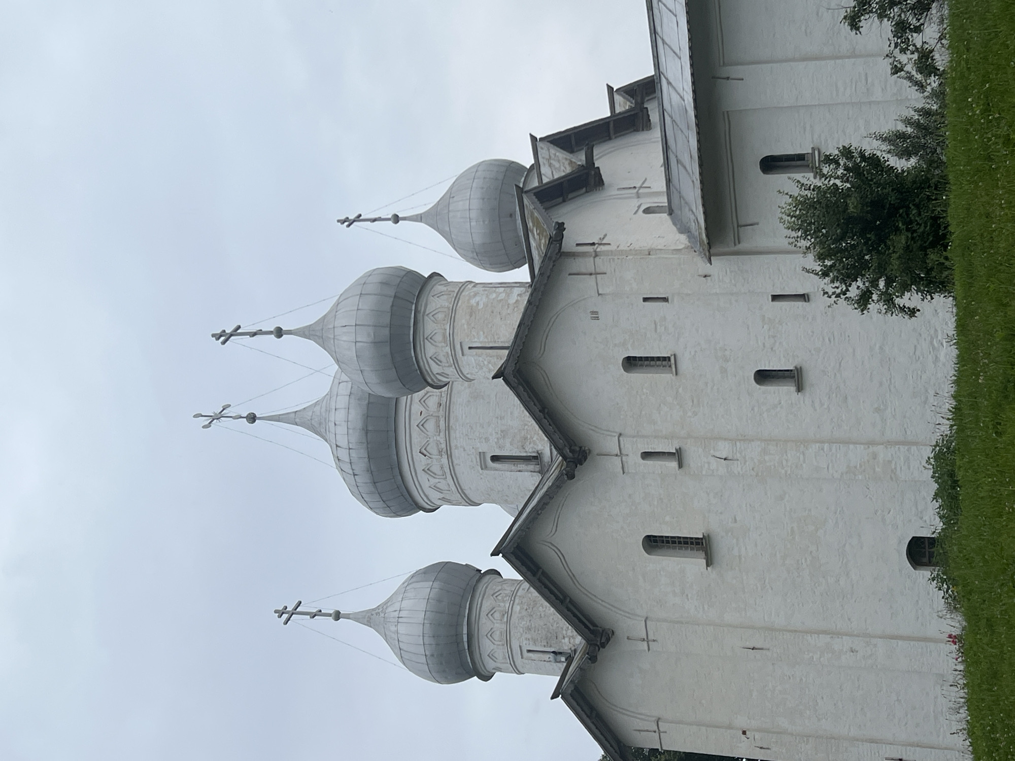 The image shows the Saint Sophia Cathedral in Veliky Novgorod, Russia. It is a white stone building with multiple silver onion domes, each topped with a cross. The cathedral has several arched windows and a pitched roof. In the foreground, there is a grassy hill with some bushes and flowers. The sky is overcast and gray.