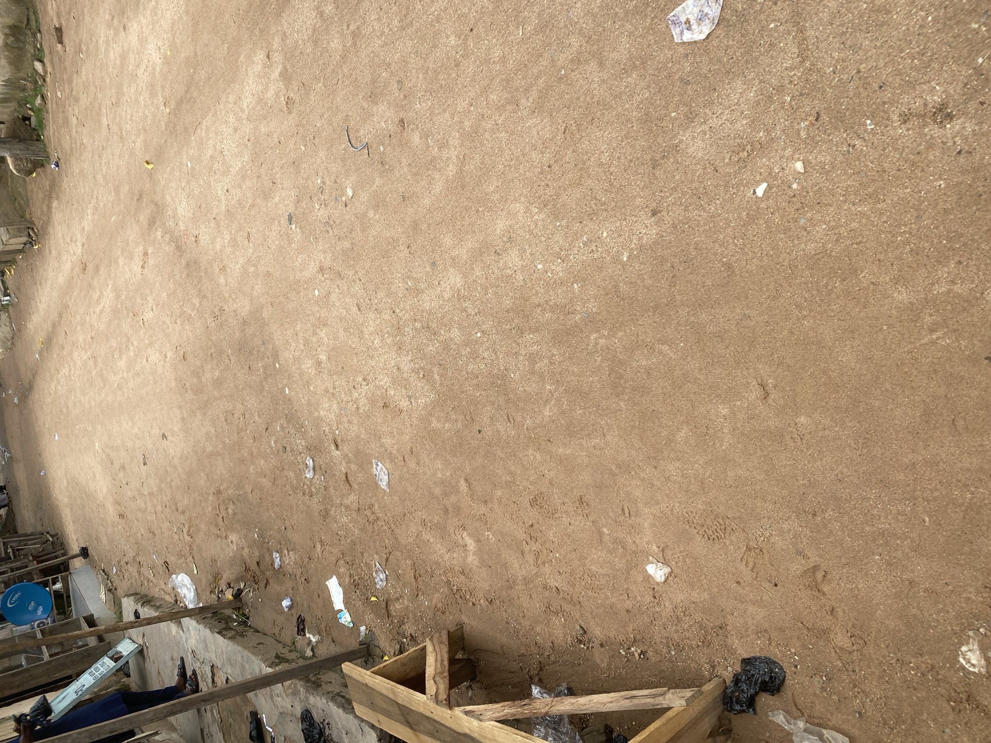 The image shows a dirt ground with scattered debris, including plastic bags and wrappers. In the lower left corner, a person is visible, wearing blue pants and sandals, sitting on a wooden structure. To their left, a blue satellite dish is mounted on a wooden frame. The scene appears to be an outdoor area, possibly a market or a street, in Jalingo, Nigeria. The ground is uneven and dusty, with some footprints visible. The lighting suggests it could be daytime. There is no discernible text in the image. The overall impression is one of a somewhat unkempt environment.