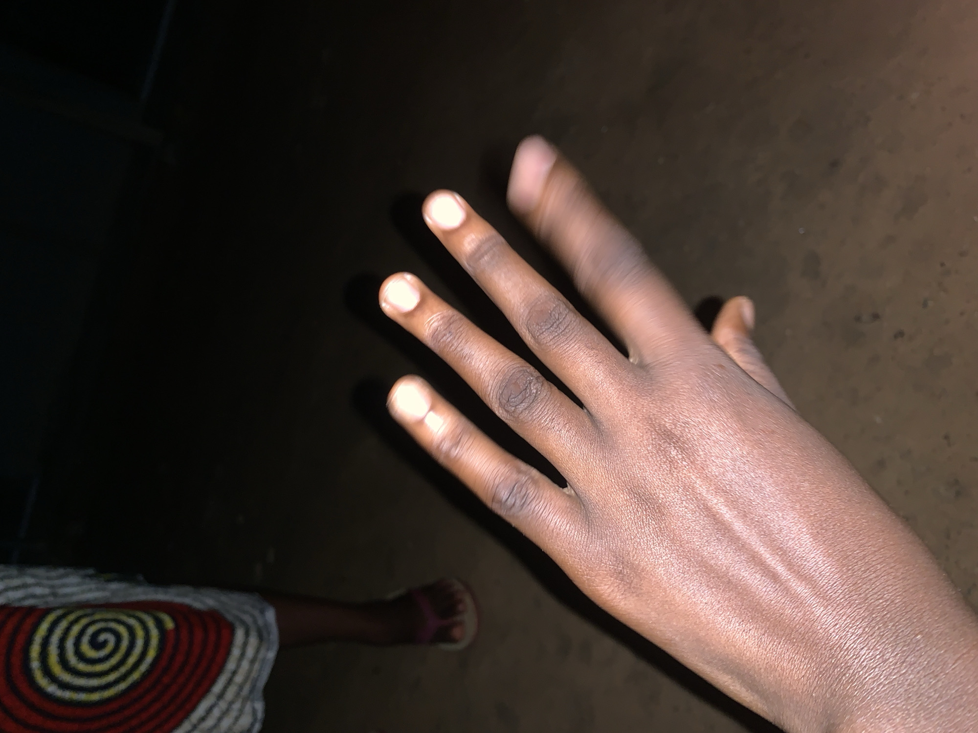 A hand is visible in the foreground against a dark background. A portion of a person's leg and foot wearing a sandal, along with a patterned skirt, are also visible in the lower left corner. The location is Keffi, Nigeria. The scene is dimly lit.