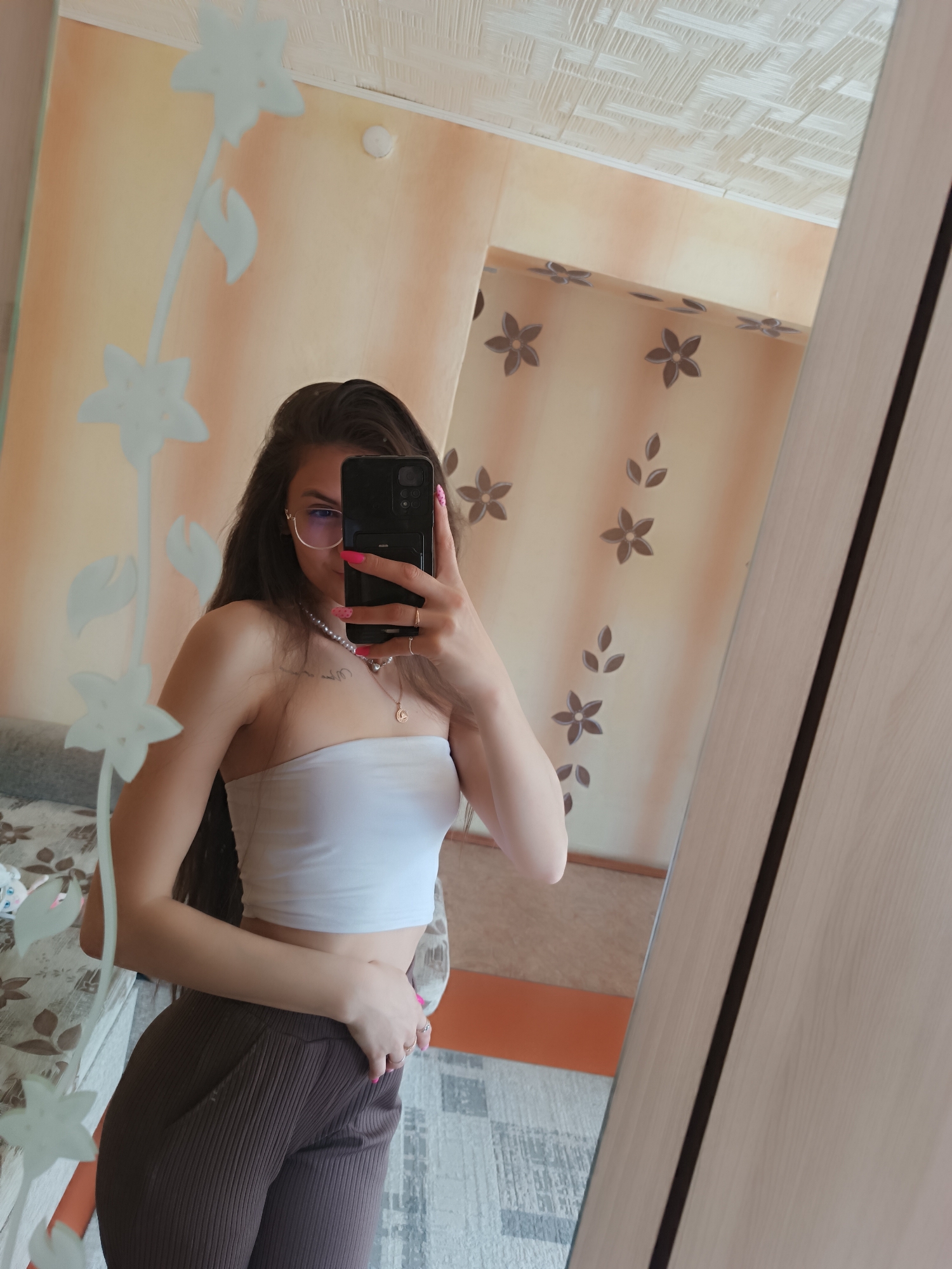 A woman takes a selfie in a mirror. The mirror is decorated with floral patterns. The woman has long dark hair, wears glasses, a white tube top, and brown pants. She holds a black smartphone in her right hand. A pearl necklace and a gold pendant necklace are visible around her neck. The room has light peach colored walls with dark brown flower decals. A section of the room reflected in the mirror shows a patterned ceiling and a light-colored piece of furniture. The location of the scene appears to be indoors.