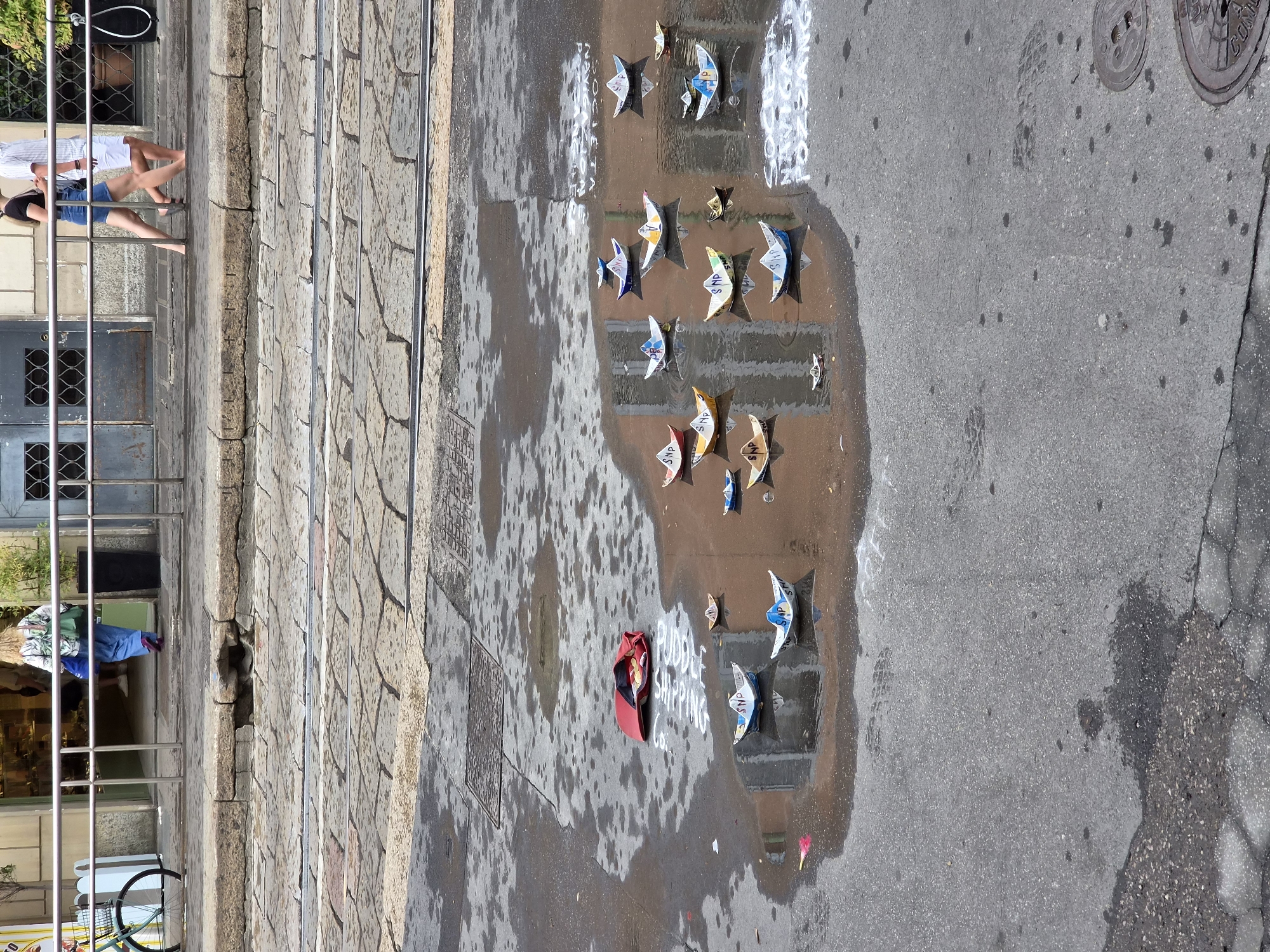 The image depicts a wet street scene in Milan, Italy, likely after rain. The foreground is dominated by a large puddle on asphalt, reflecting the surroundings. Several origami paper boats are floating in the puddle, some decorated with white lettering, possibly political slogans. A red bag lies open near the edge of the puddle. In the background, cobblestone pavement and tram tracks are visible. Beyond that, a sidewalk with a metal railing separates the street from a building facade. People are walking on the sidewalk, with a woman in blue pants and a patterned shirt in the process of entering a store, and two individuals, a man and a woman, walking barefoot. A bicycle with a basket is parked on the left side of the image.