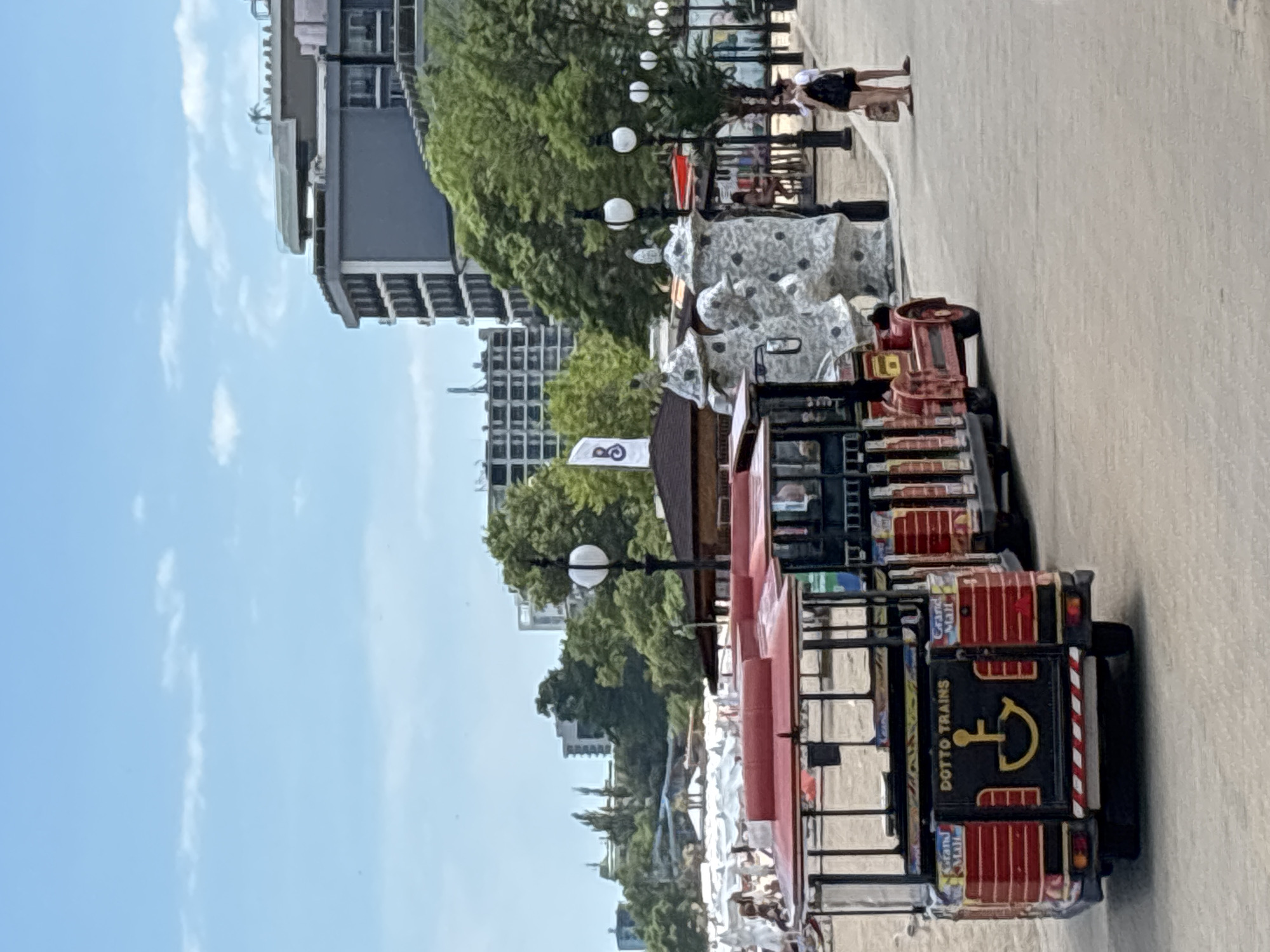 This is a vertically oriented, full shot of a street scene in Varna, Bulgaria. In the foreground, a red and black "Dotto Trains" vehicle, a type of trackless train, is parked. The vehicle has a black rear with red striped accents, a yellow anchor symbol, and the text "DOTTO TRAINS" written on it. Behind the train, there are several white structures that appear to be part of a playground or activity area.
Further back, tall, modern buildings with many windows are visible, interspersed with lush green trees. A street lamp with a white globe is positioned among the trees. The sky is a bright blue with scattered white clouds, suggesting a sunny daytime. Two people are standing on the paved pathway in the middle ground, with one person, likely a child wearing a dark dress and white hat, facing away from the camera and walking. The overall scene appears to be a public area, possibly a promenade or park, on a clear day.