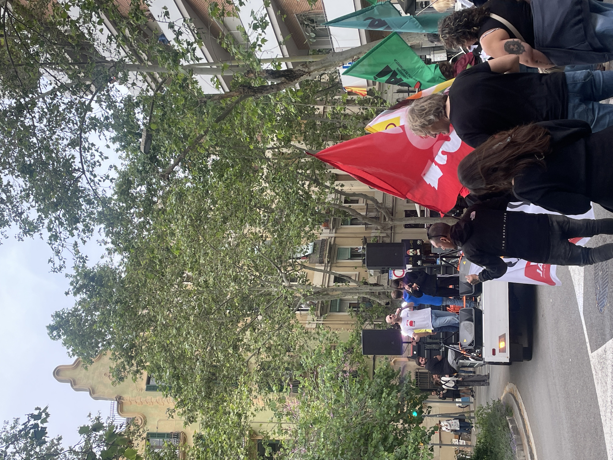 This image depicts an outdoor public gathering, likely a rally or demonstration, taking place on a city street. A white truck serves as a temporary stage, equipped with large black speakers. Several individuals are on or around this stage; one person in a white T-shirt with a red graphic appears to be speaking into a microphone, while others stand nearby.
A crowd of people is gathered, many holding various flags. Prominent among these are a large red flag bearing a distinctive white stylized symbol (resembling an inverted V with a circle on top) and several green flags. Text partially visible on one green flag includes "PAÍS VALENCIÀ," suggesting a connection to Valencian regional identity or political movements. Another flag, yellow and orange with a visible symbol, is also present.
The scene is set on a tree-lined street, with mature trees partially obscuring multi-story buildings in the background. One building on the left side features ornate architectural details, indicative of a Modernista or Art Nouveau style, consistent with structures found in Barcelona. The lighting suggests it is daytime, possibly under an overcast sky, as shadows are soft and diffused. The event conveys an atmosphere of public engagement.